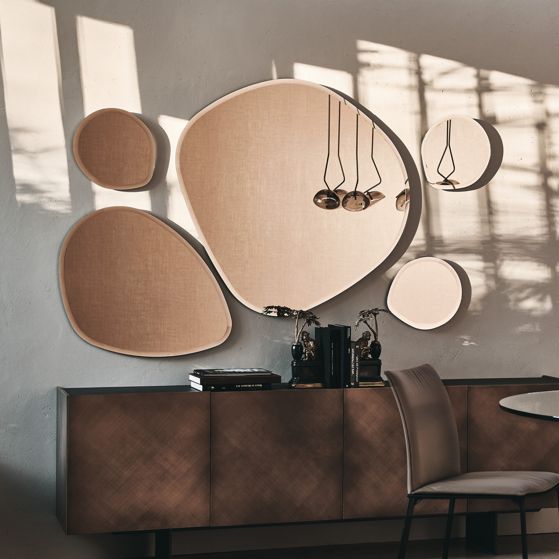 A modern interior with a grey wall adorned with five irregularly shaped, beige and mirrored panels, casting shadows. Below is a dark brown credenza with books and monkey figurines. A taupe chair and table are visible on the right.