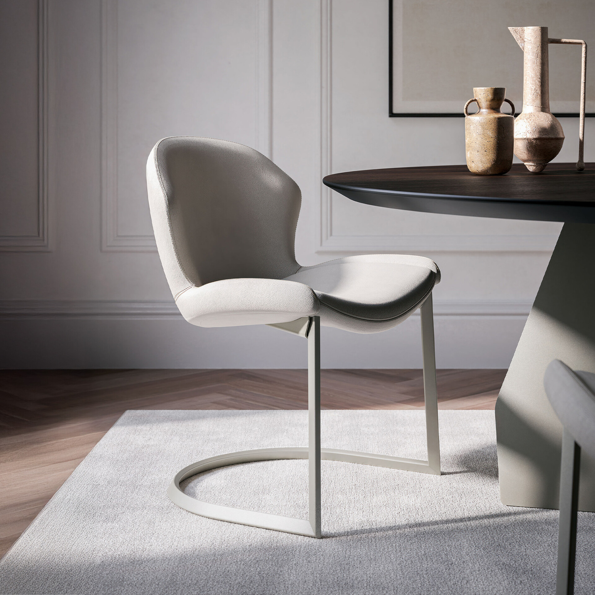 A modern light grey upholstered dining chair with a unique cantilever base sits on a light rug next to a dark wood dining table adorned with rustic vases, in a contemporary room with paneled walls.