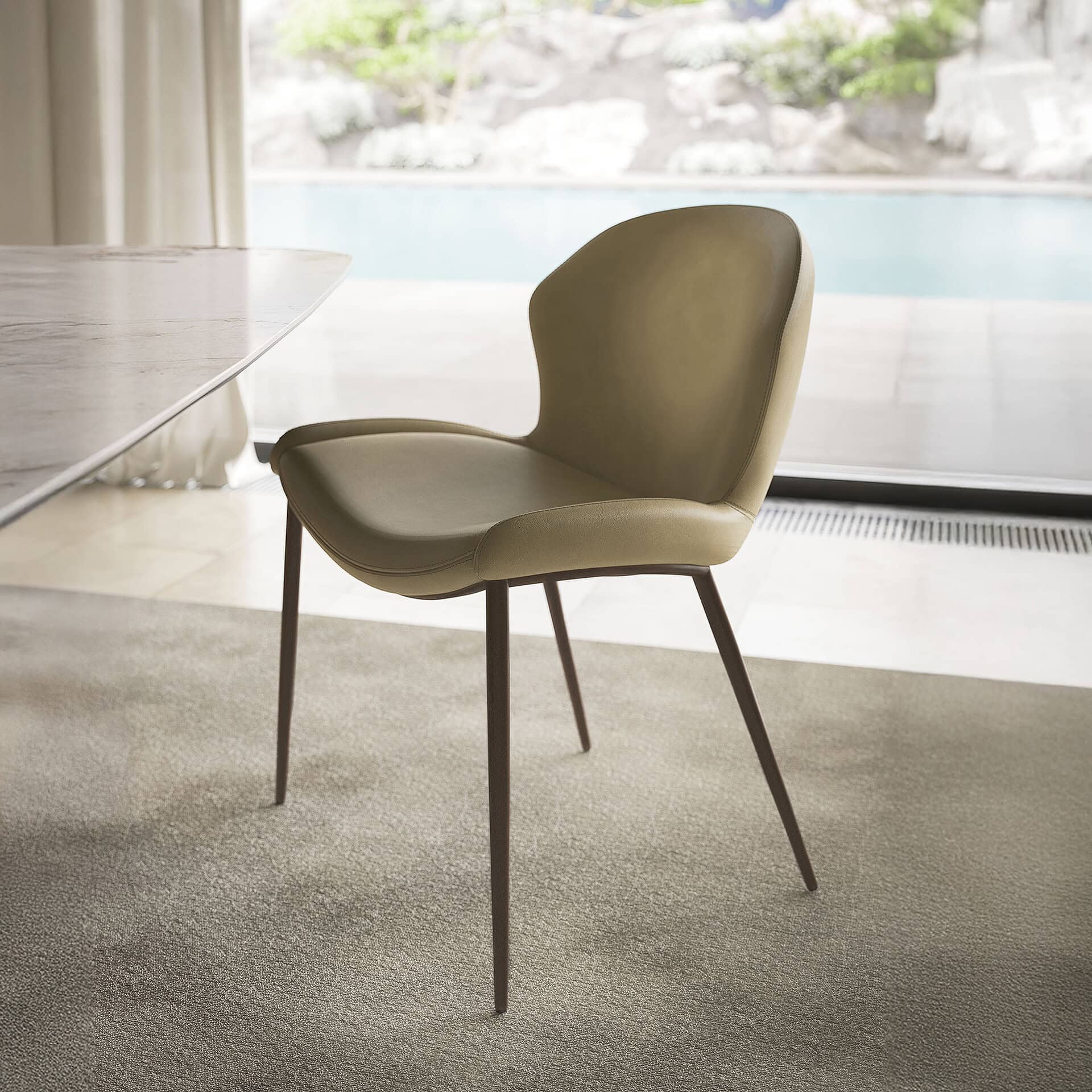 A modern, light olive green leather dining chair with dark metal legs sits on a beige carpet, next to a marble table, overlooking a swimming pool and lush garden through a large window.