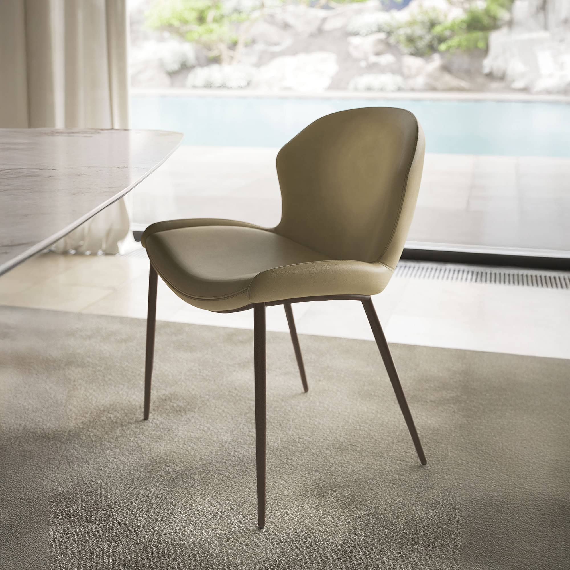 A modern, light olive green leather dining chair with dark metal legs sits on a beige carpet, next to a marble table, overlooking a swimming pool and lush garden through a large window.