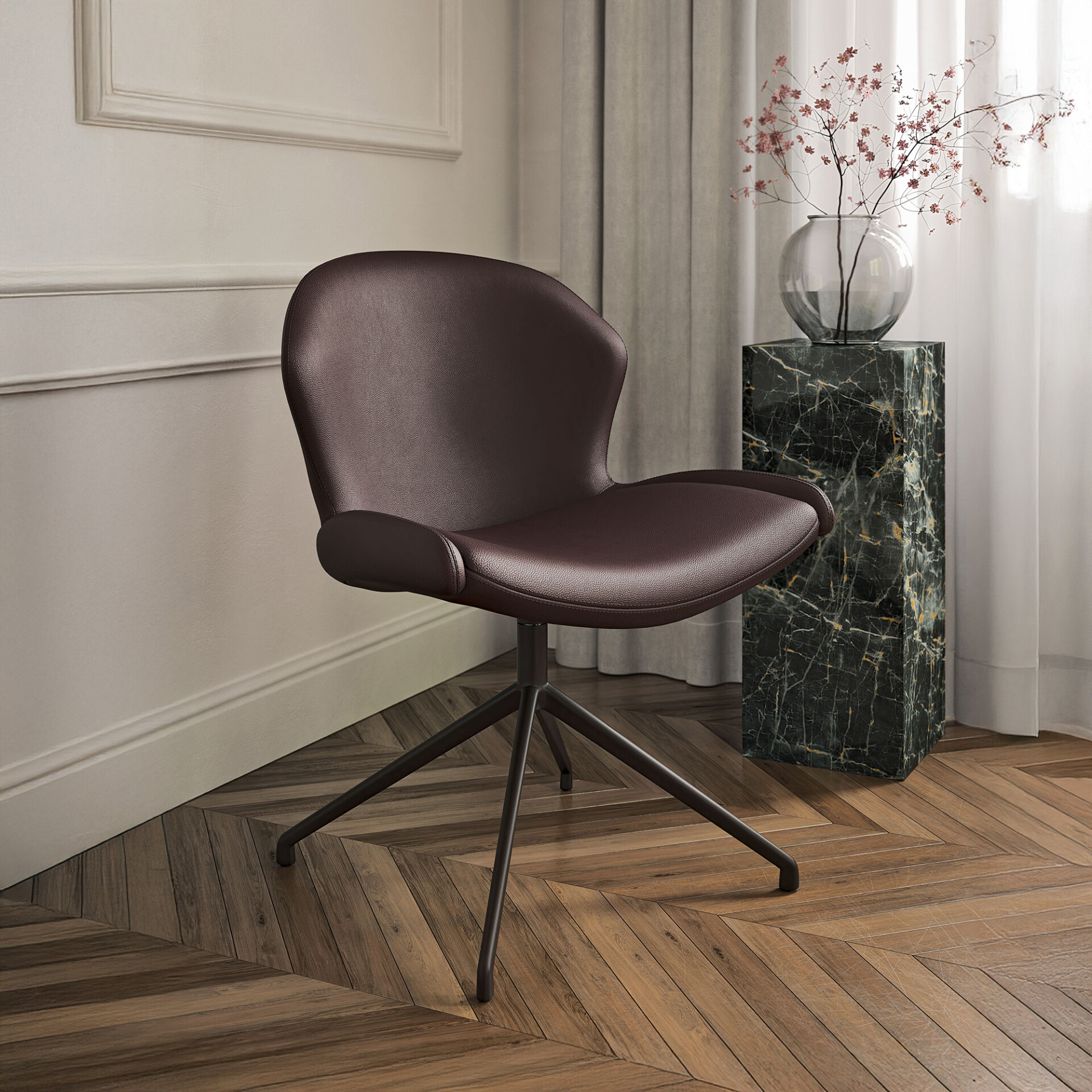 A modern dark brown leather chair with a dark metal base sits on a herringbone wood floor. Behind it, a green marble pedestal holds a clear vase with pink blossoms, next to a window with sheer curtains and a light wall with molding.