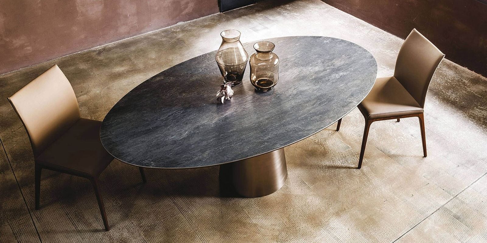An overhead view of a modern oval dining table with a dark, textured top and a bronze pedestal base. Two tan leather chairs are placed on either side. Two amber glass vases and a small decorative object sit on the table. The room features a light textured rug and a muted reddish-brown wall. A Cattelan Yoda Dining Table.