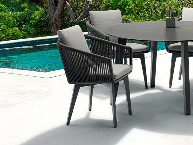 Modern dark grey outdoor dining chairs and table with light grey cushions on a white patio next to a turquoise swimming pool, surrounded by green foliage.