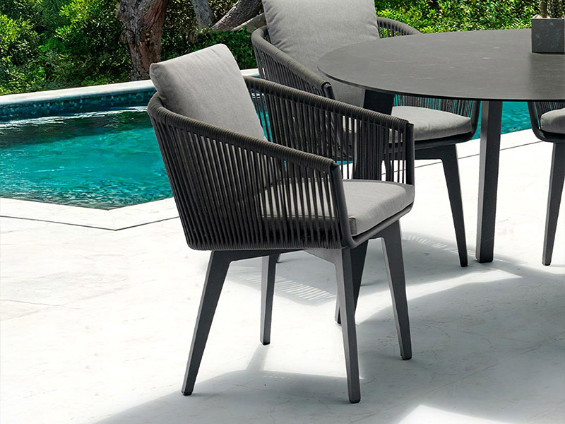 Modern dark grey outdoor dining chairs and table with light grey cushions on a white patio next to a turquoise swimming pool, surrounded by green foliage.