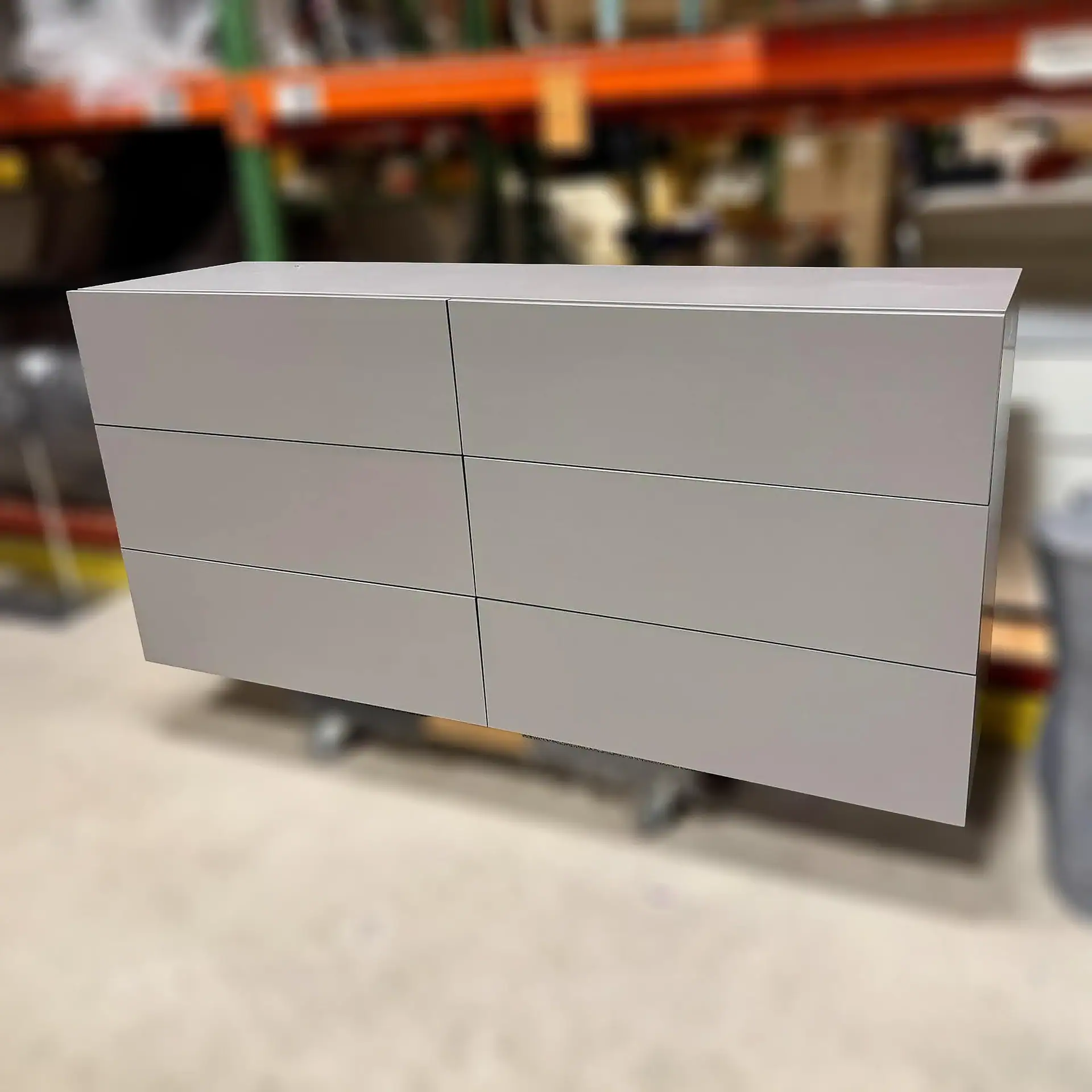 Image shows a sleek, minimalist gray dresser with six spacious drawers in a furniture warehouse.