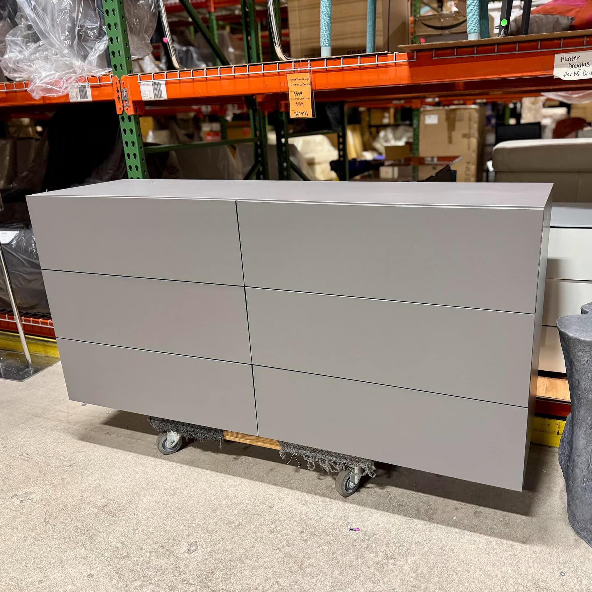 Image shows a sleek, minimalist gray dresser with six spacious drawers in a furniture warehouse.