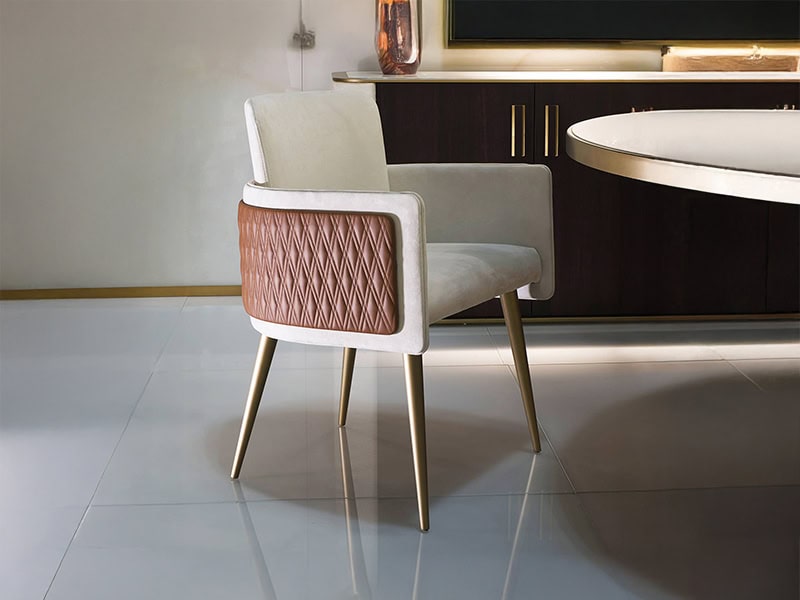 A modern cream upholstered chair with a quilted brown leather back and gold legs, positioned in an elegant dining room setting with a dark cabinet and round table in the background.