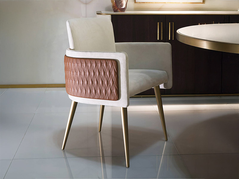 A modern cream upholstered chair with a quilted brown leather back and gold legs, positioned in an elegant dining room setting with a dark cabinet and round table in the background.