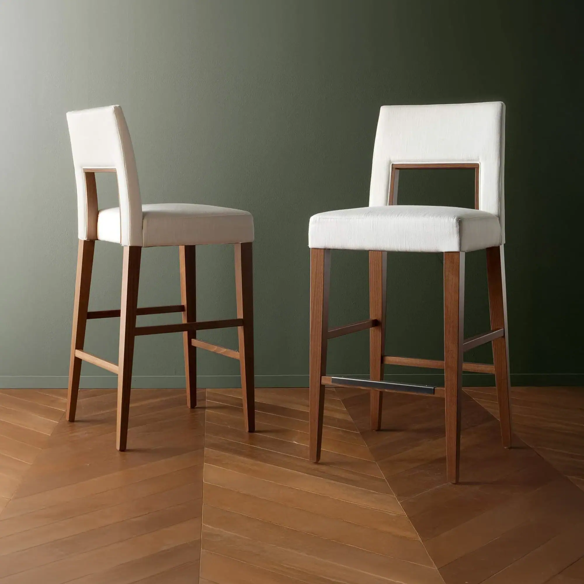 Two modern bar stools with light fabric upholstery and dark wooden frames stand on a herringbone wood floor against a green wall.