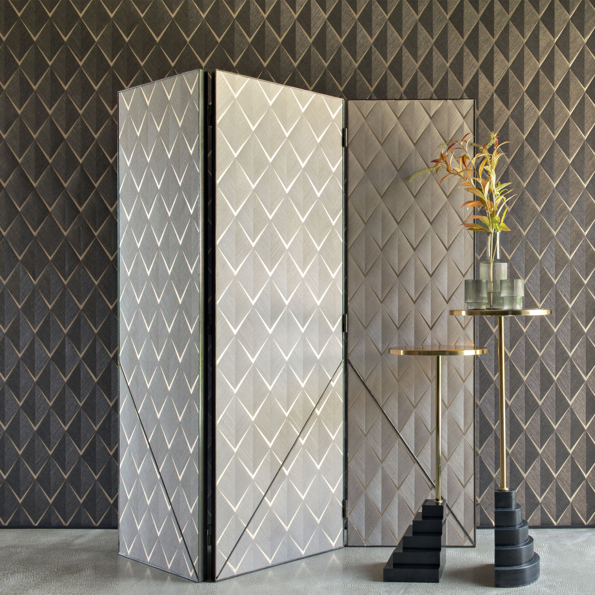 A luxurious room with a dark geometric patterned wall, a light grey three-panel room divider with matching patterns, and a brass side table with a black sculptural base holding a vase of autumn foliage.