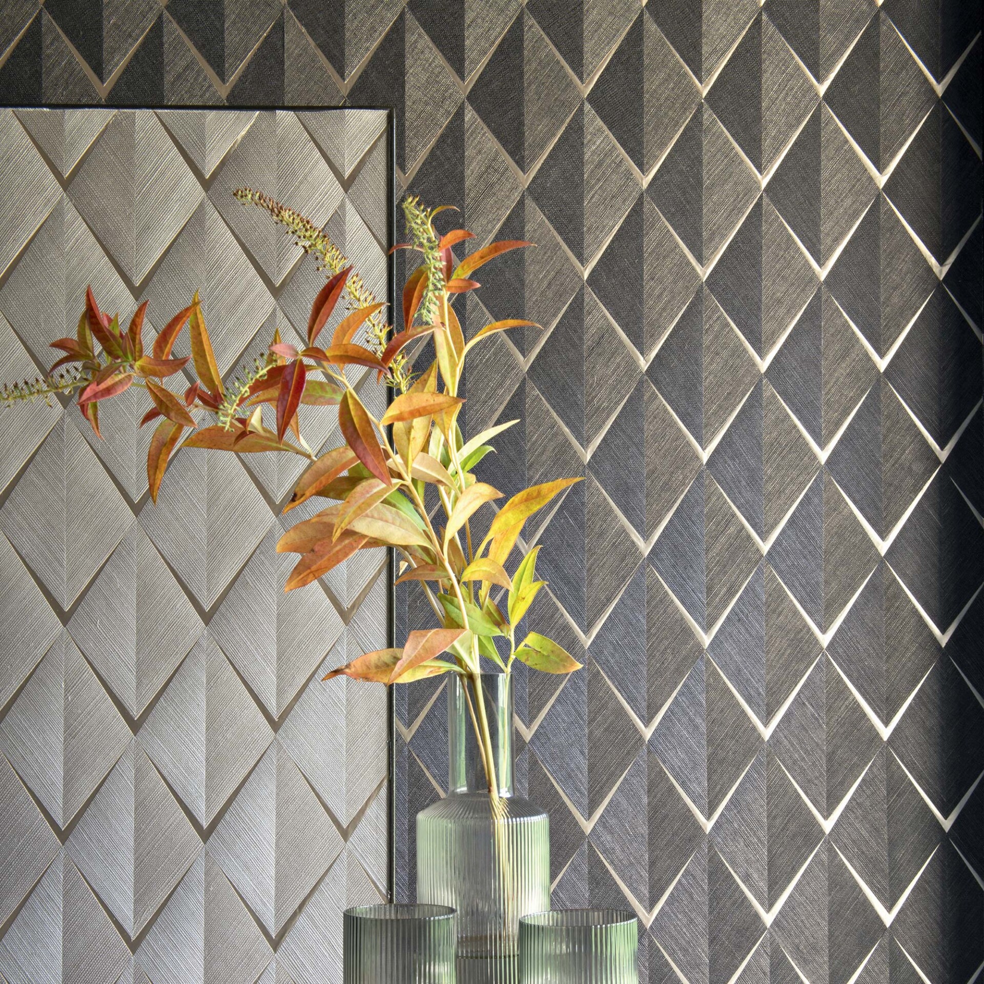 A clear fluted glass vase filled with green, yellow, and orange autumnal leaves sits in front of a textured wall. The wall features an embossed geometric diamond pattern, with lighter gray diamonds on the left and darker gray diamonds on the right, separated by a dark vertical frame.
