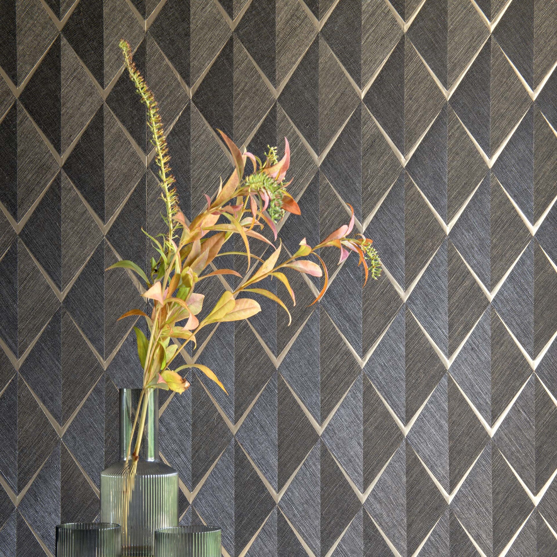 A clear, ribbed glass vase with artificial foliage in muted green, yellow, and orange tones, set against a dark geometric wallpaper. The wallpaper features a repeating diamond pattern with textured, faceted details and light-colored outlines.
