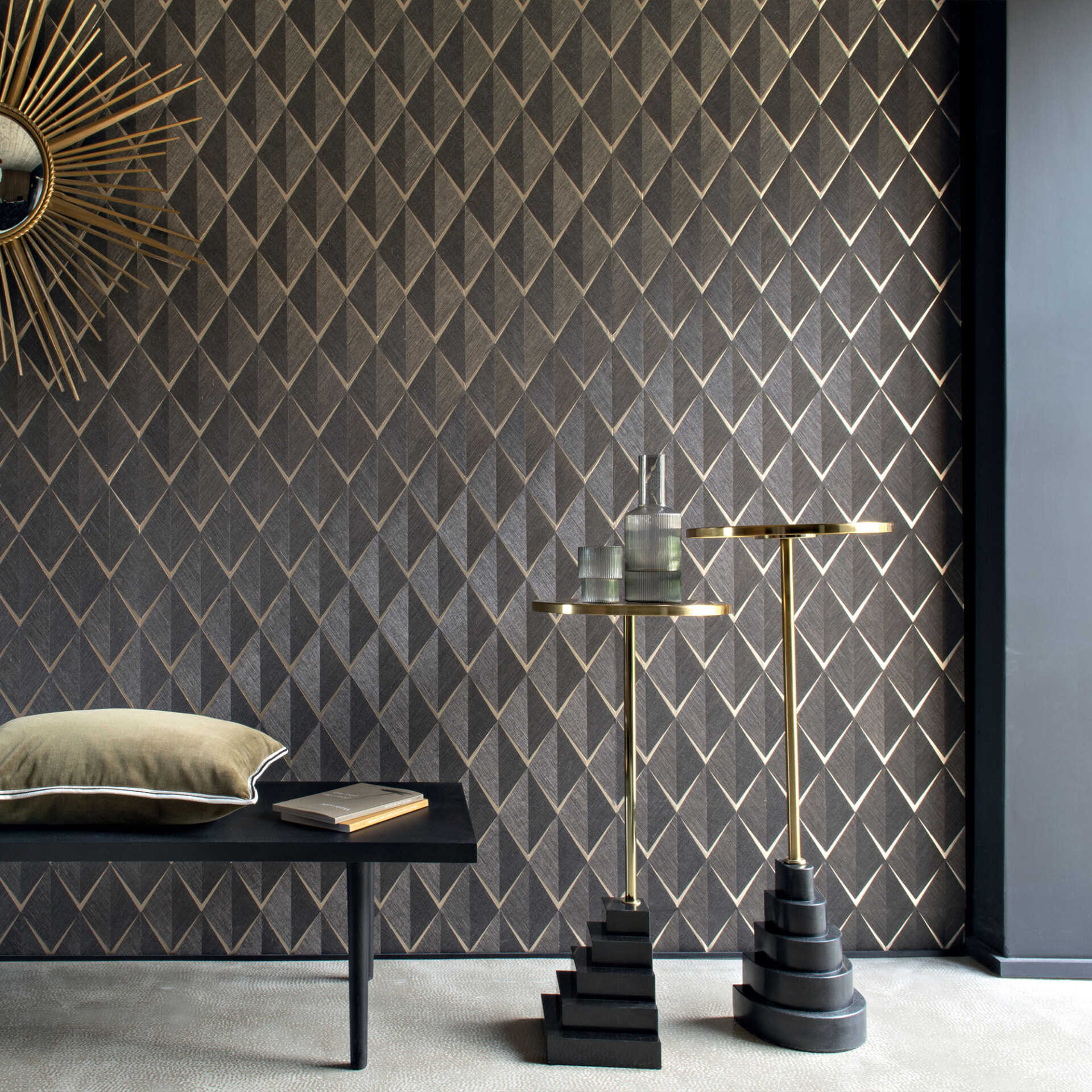 An elegant room featuring dark geometric wallpaper with gold accents, a gold sunburst mirror, a black bench with a green cushion, and two modern gold side tables with glass decor.