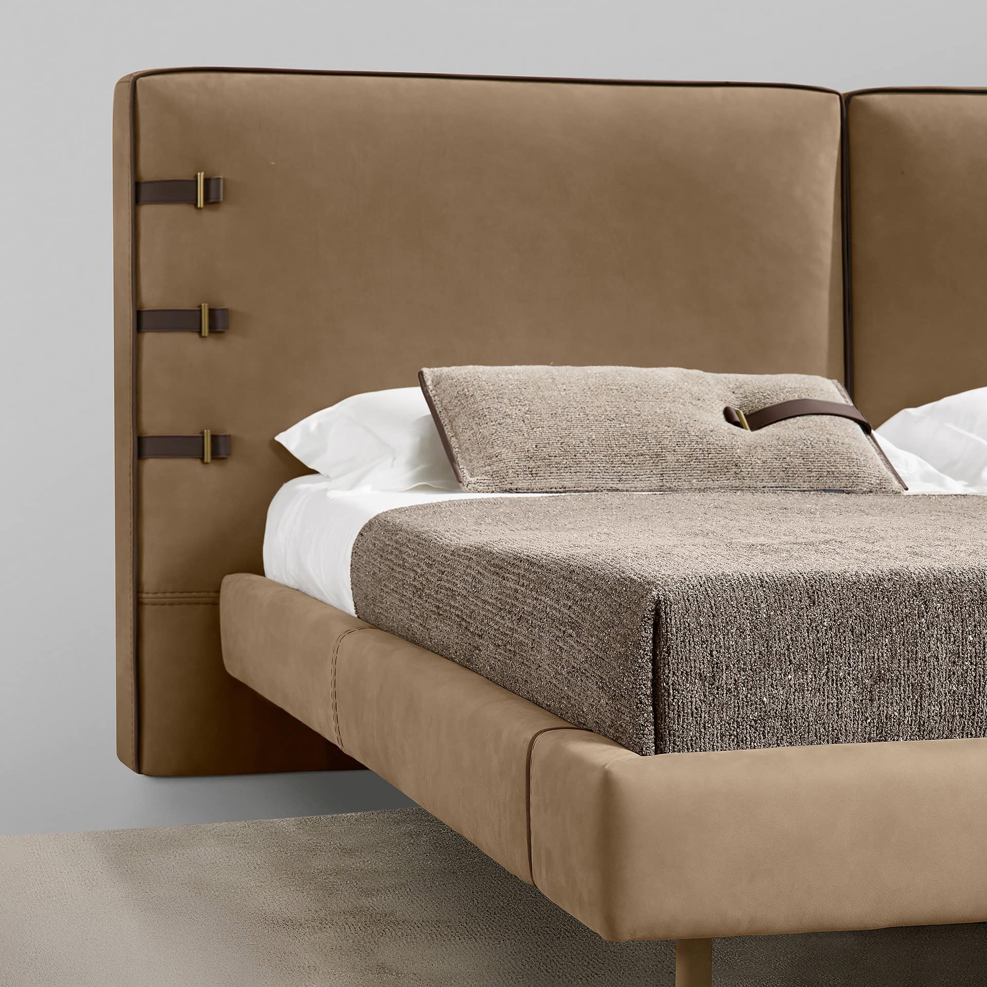 A modern bed with a tan leather headboard featuring dark brown straps and brass buckles. The bed has textured taupe bedding and white sheets.