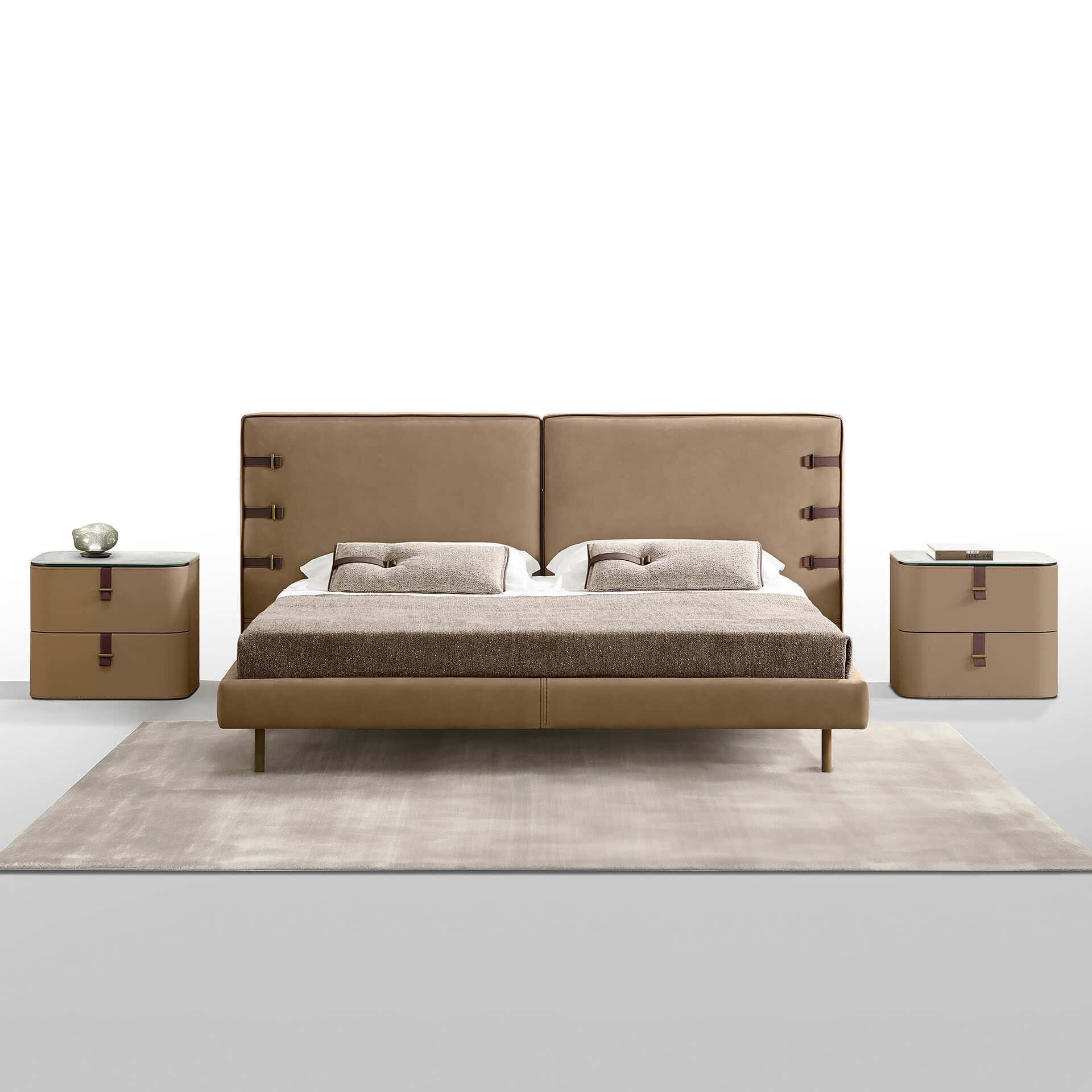 A modern bedroom set with a tan leather bed and two matching nightstands on a light beige rug. The bed has a headboard with dark brown leather straps and brass buckles. The nightstands also feature matching strap and buckle drawer pulls. A decorative object is on the left nightstand, and a book is on the right.
