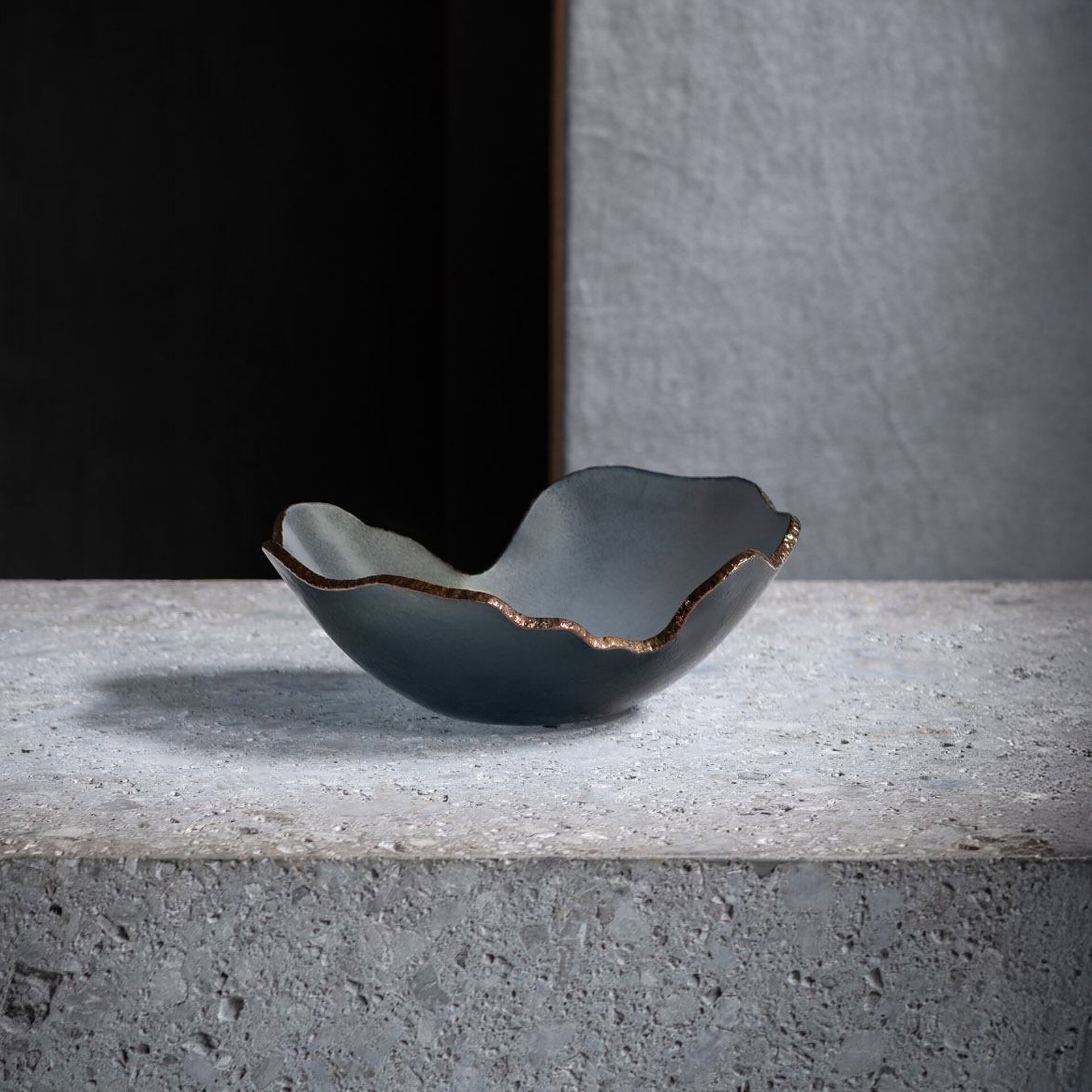 A blue-gray, wavy-edged bowl with a gold rim sits on a gray concrete surface against a split dark and light gray background.