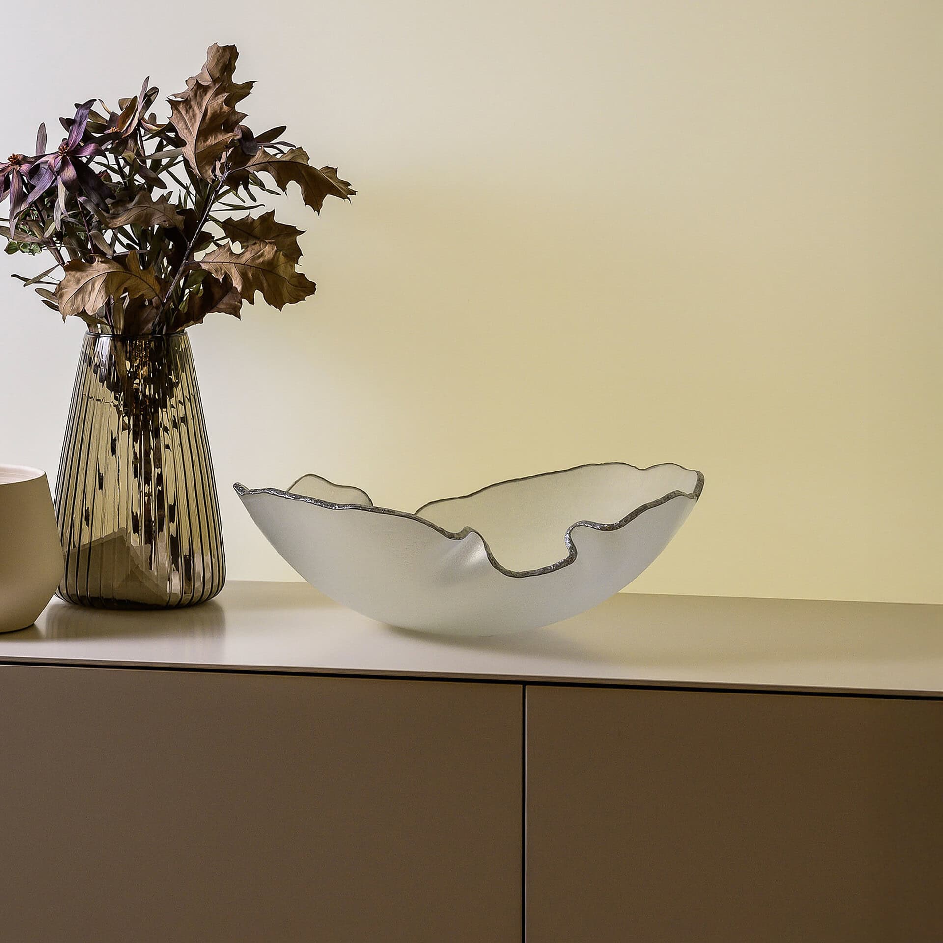 A decorative display on a modern cabinet featuring a dark fluted glass vase filled with dried brown and purple leaves, and a frosted white glass bowl with a wavy silver-edged rim, against a pale yellow wall.