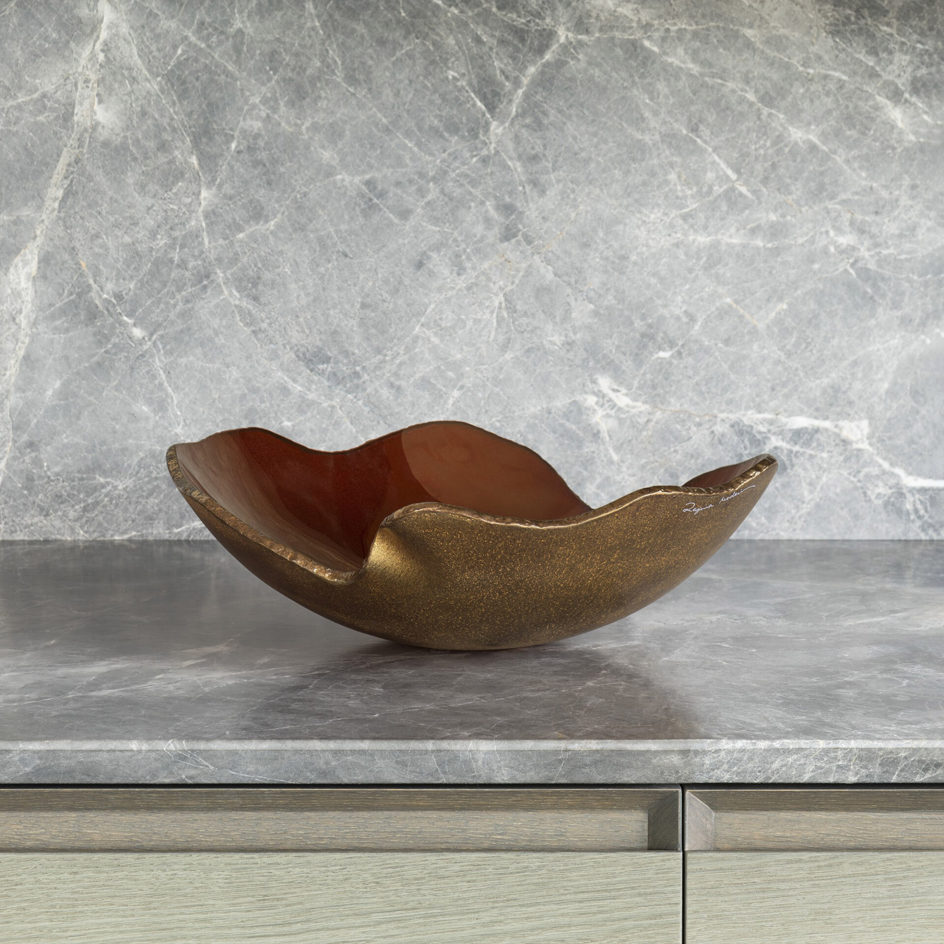 A decorative, organically shaped bowl with a textured bronze exterior and glossy reddish-brown interior, resting on a gray marble countertop with a matching backsplash. Light wood cabinetry is visible below.