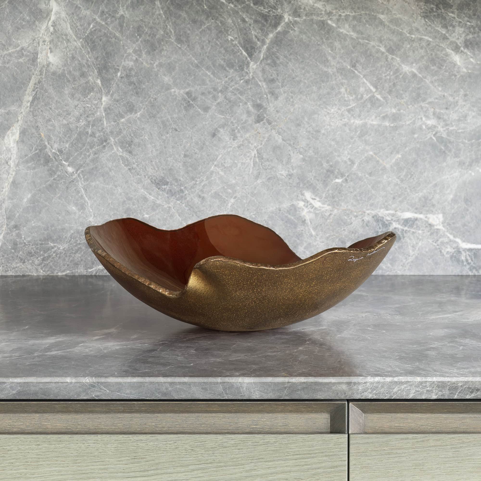 A decorative, organically shaped bowl with a textured bronze exterior and glossy reddish-brown interior, resting on a gray marble countertop with a matching backsplash. Light wood cabinetry is visible below.