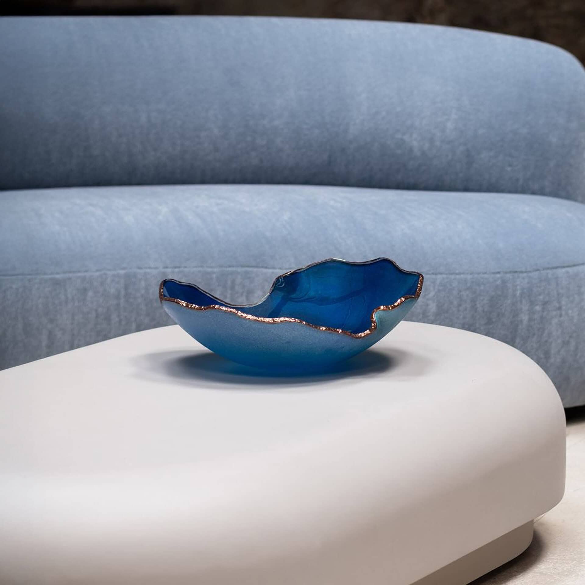 A decorative bowl with a deep blue interior and lighter blue exterior, edged in coppery-gold, sits on a light grey modern coffee table. A blurred light blue sofa is in the background.