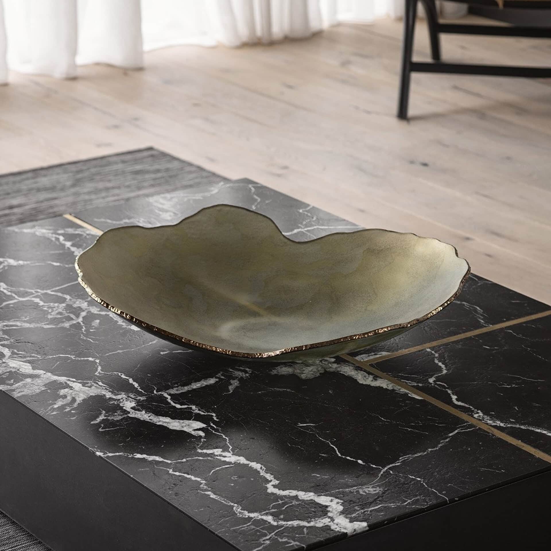 An irregular, olive green decorative bowl with a gold rim sits on a black marble coffee table with white veining and gold inlays. Light wooden floor and white curtains are in the background.