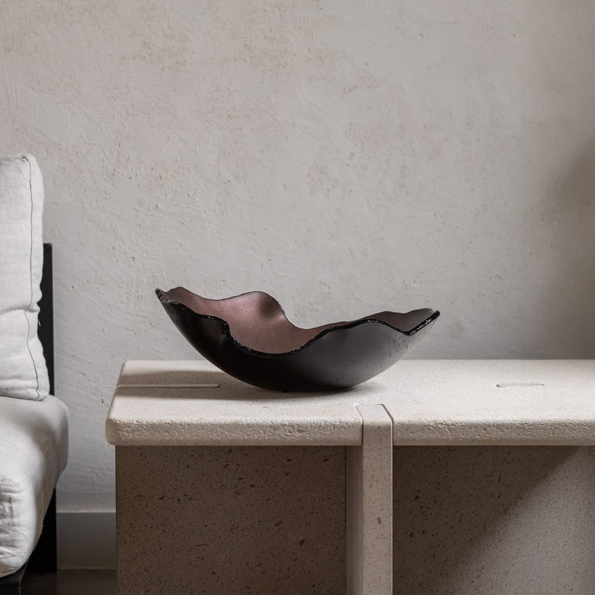 A dark, wavy decorative bowl with a metallic copper interior sits on a light, textured stone table in a modern setting.