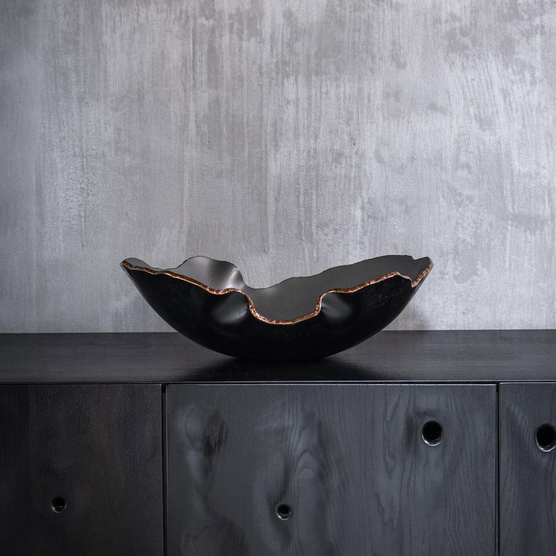 A black, irregularly shaped decorative bowl with a coppery metallic rim sits on a dark wooden cabinet against a gray textured wall.