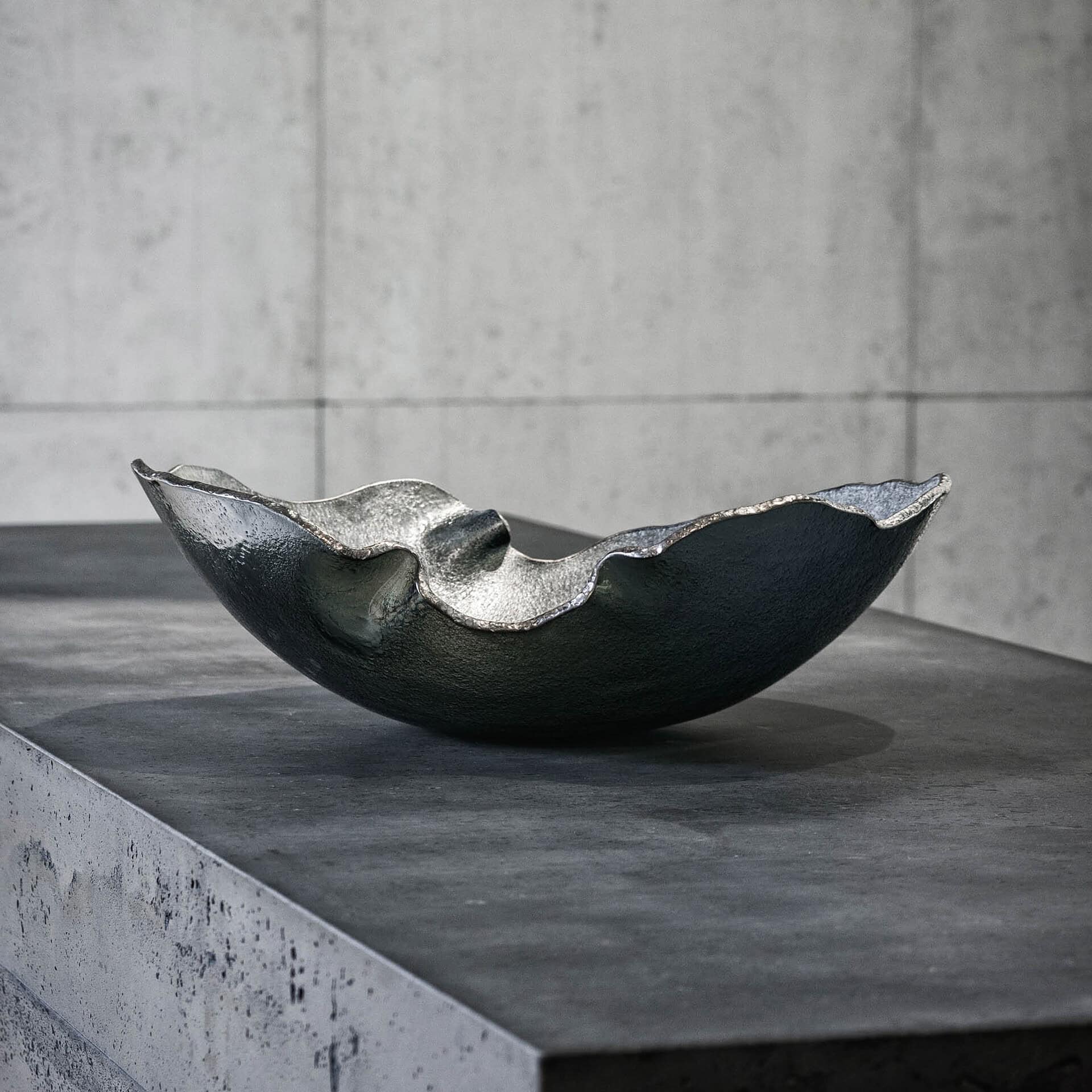 A decorative bowl with a dark, textured exterior and a shimmering, hammered silver interior and wavy rim, resting on a dark concrete surface with a lighter concrete wall in the background.