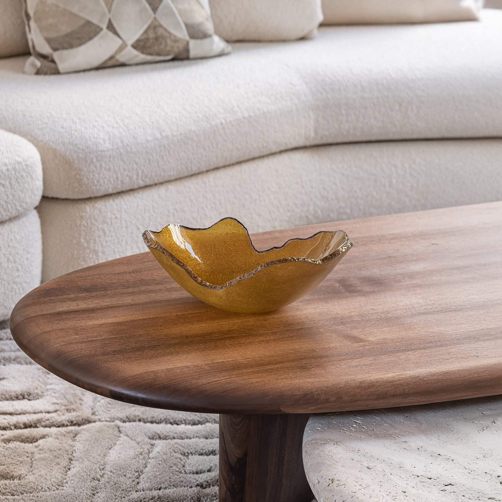 A modern living room scene with a dark wood oval coffee table, an amber glass decorative bowl with a wavy gold rim, and a cream bouclé sofa with a patterned pillow in the background.