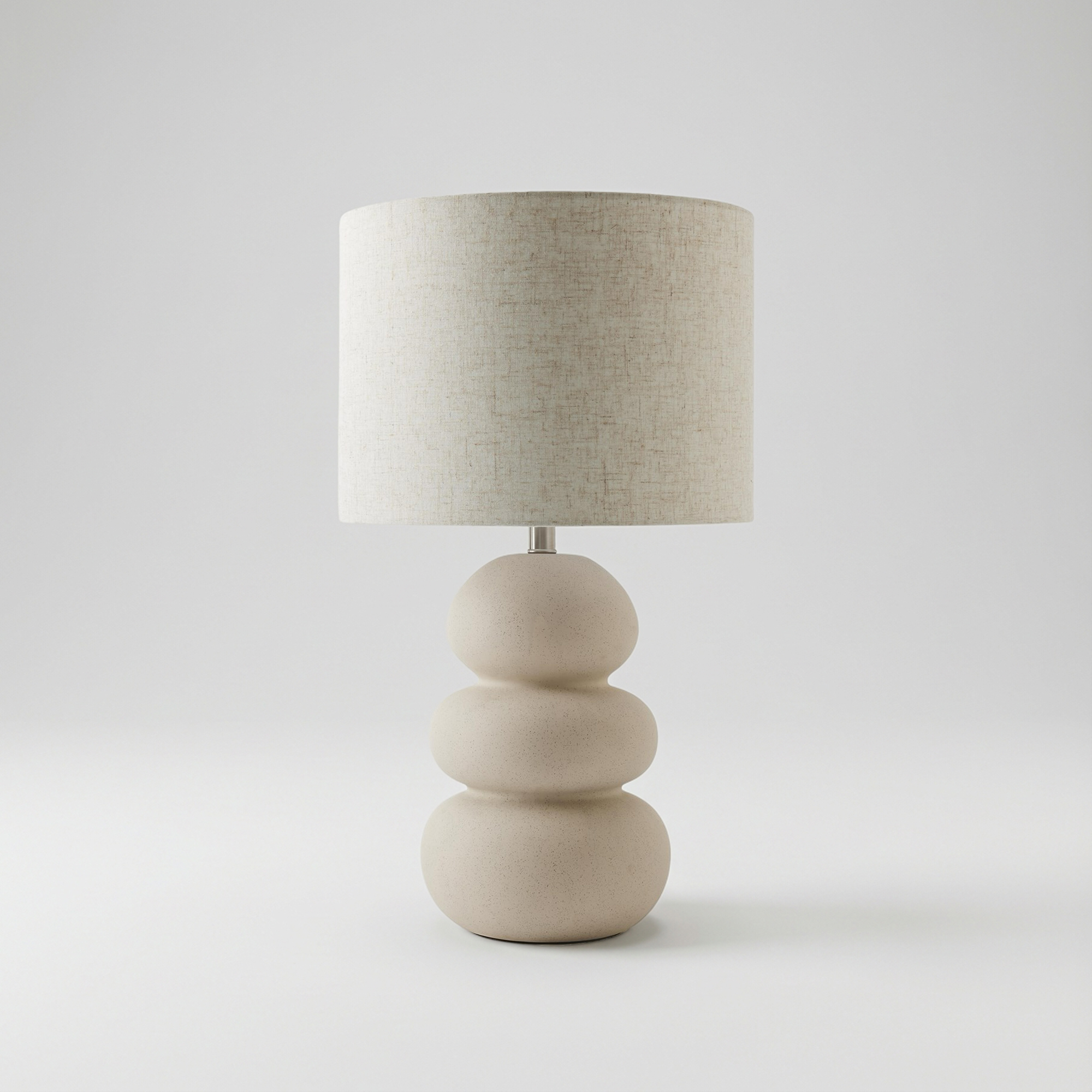 A modern table lamp with a stacked, rounded, light beige ceramic base and a textured natural linen drum shade, on a light gray background.