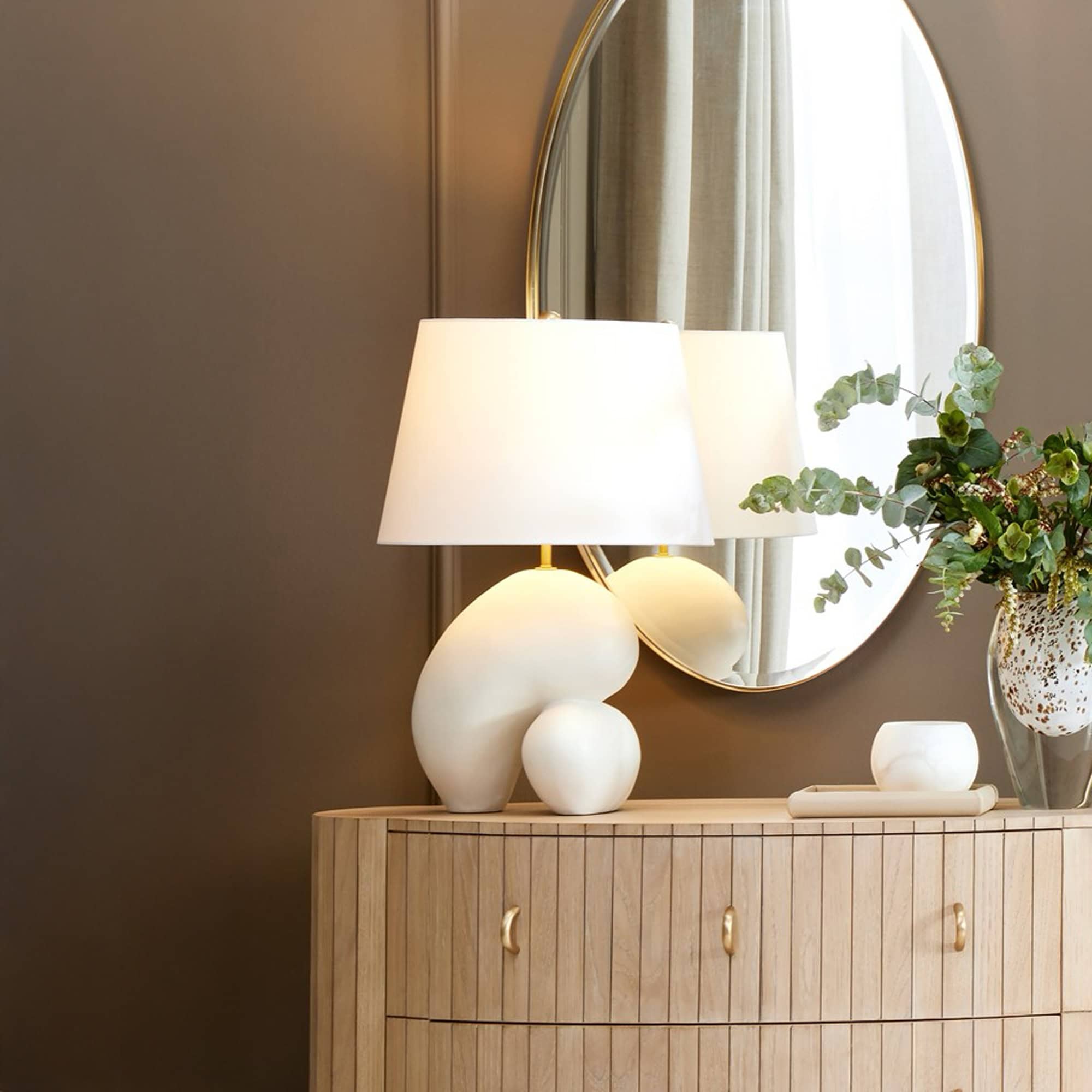 A modern interior scene with a light wood fluted dresser, a white organic-shaped table lamp, an oval gold-framed mirror, and a vase of eucalyptus, all against a brown wall.