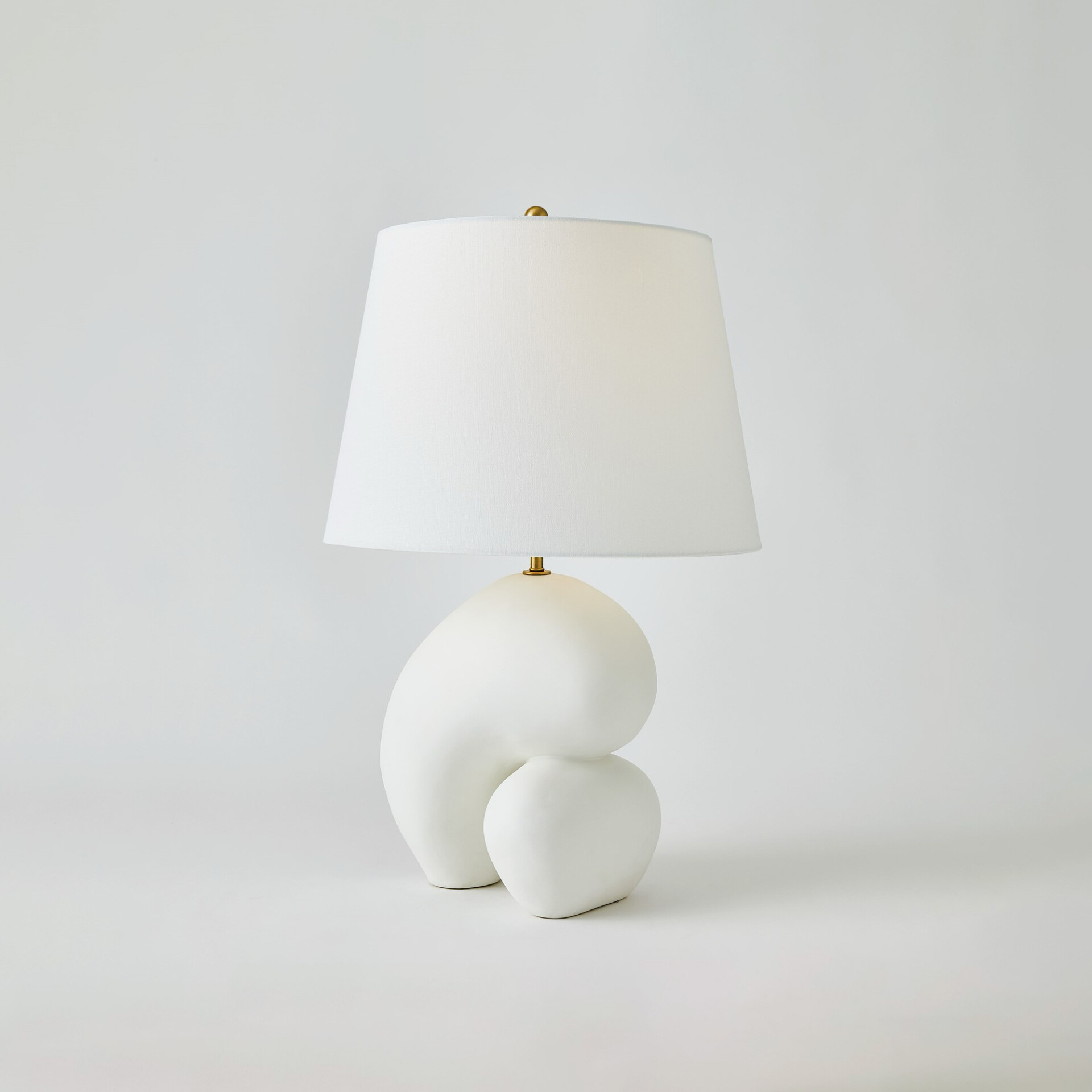 A modern table lamp with a white tapered fabric shade and a sculptural, organic matte white base, set against a light grey background.