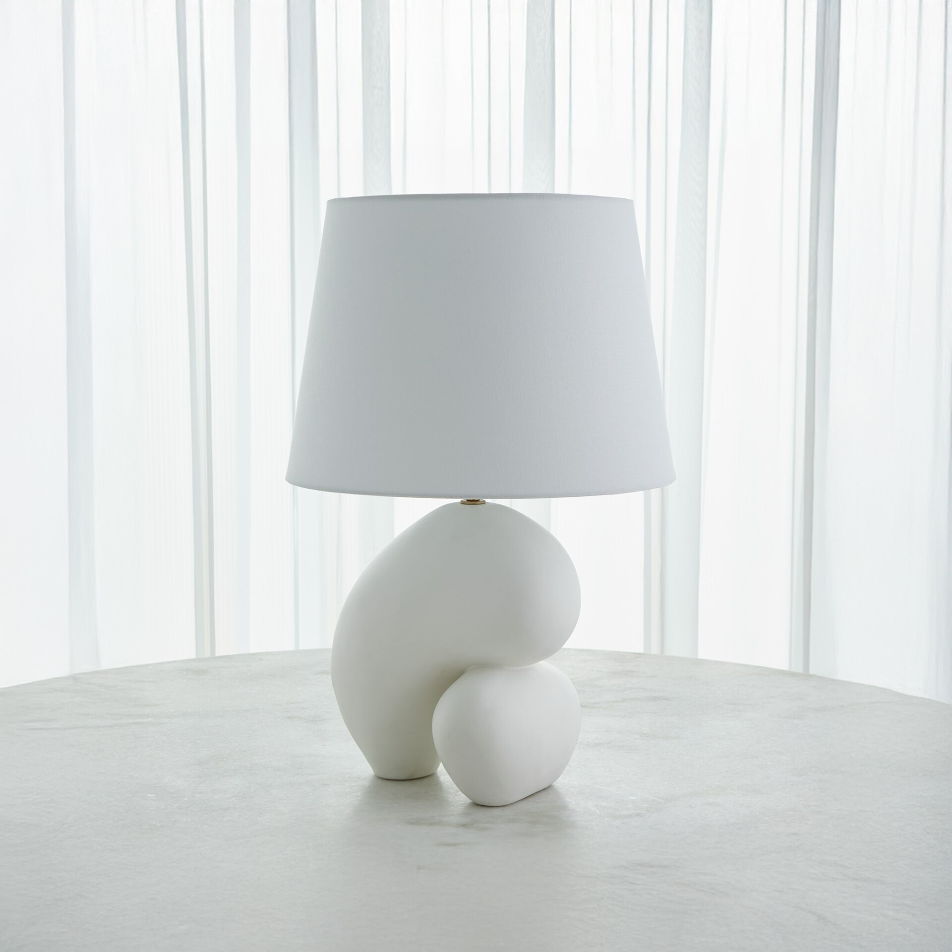 A modern white table lamp with a sculptural, organic base and a conical white shade sits on a light grey table in front of sheer white curtains.