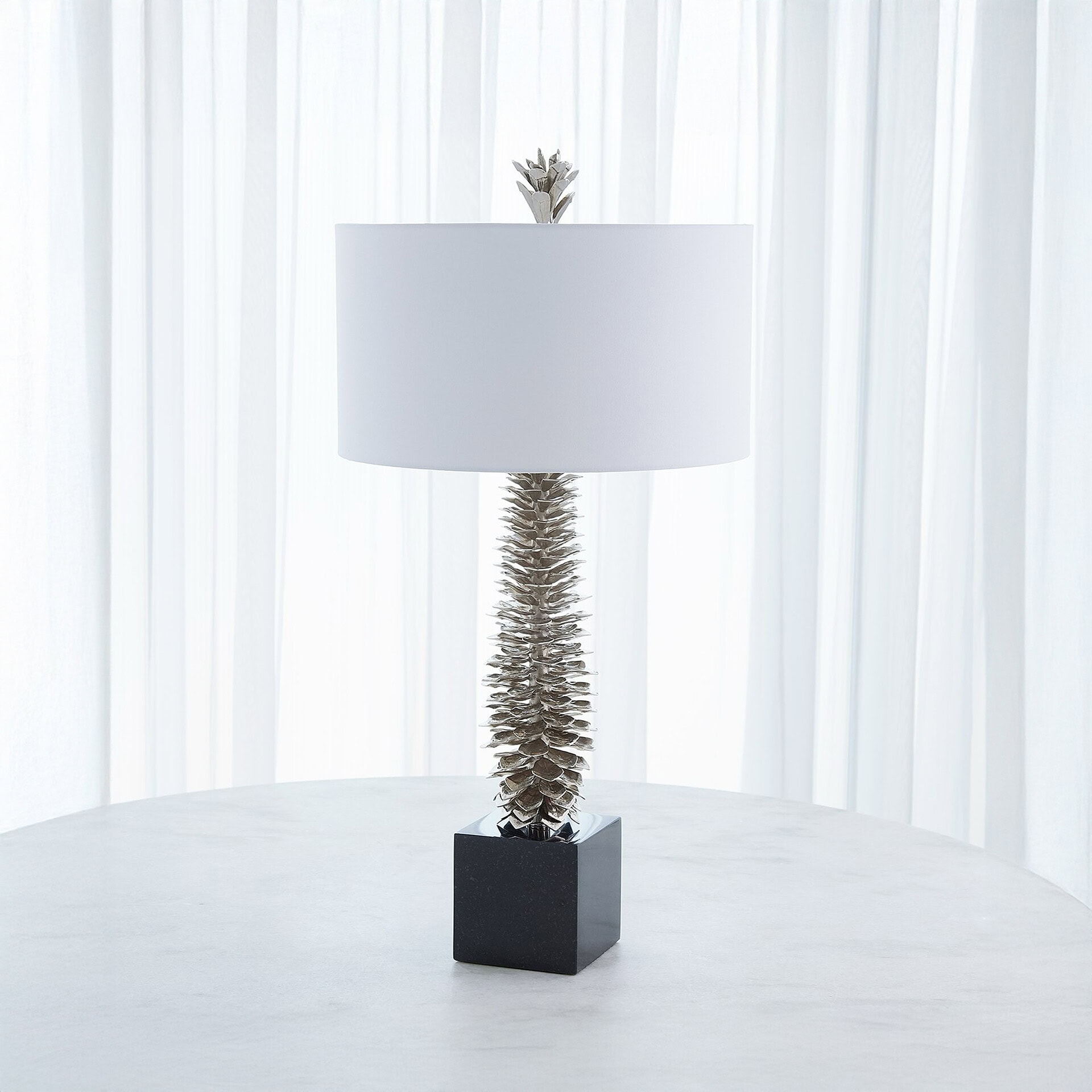 A modern table lamp with a textured silver column, white drum shade, and black cubic base, sitting on a light grey marble table in front of sheer white curtains.
