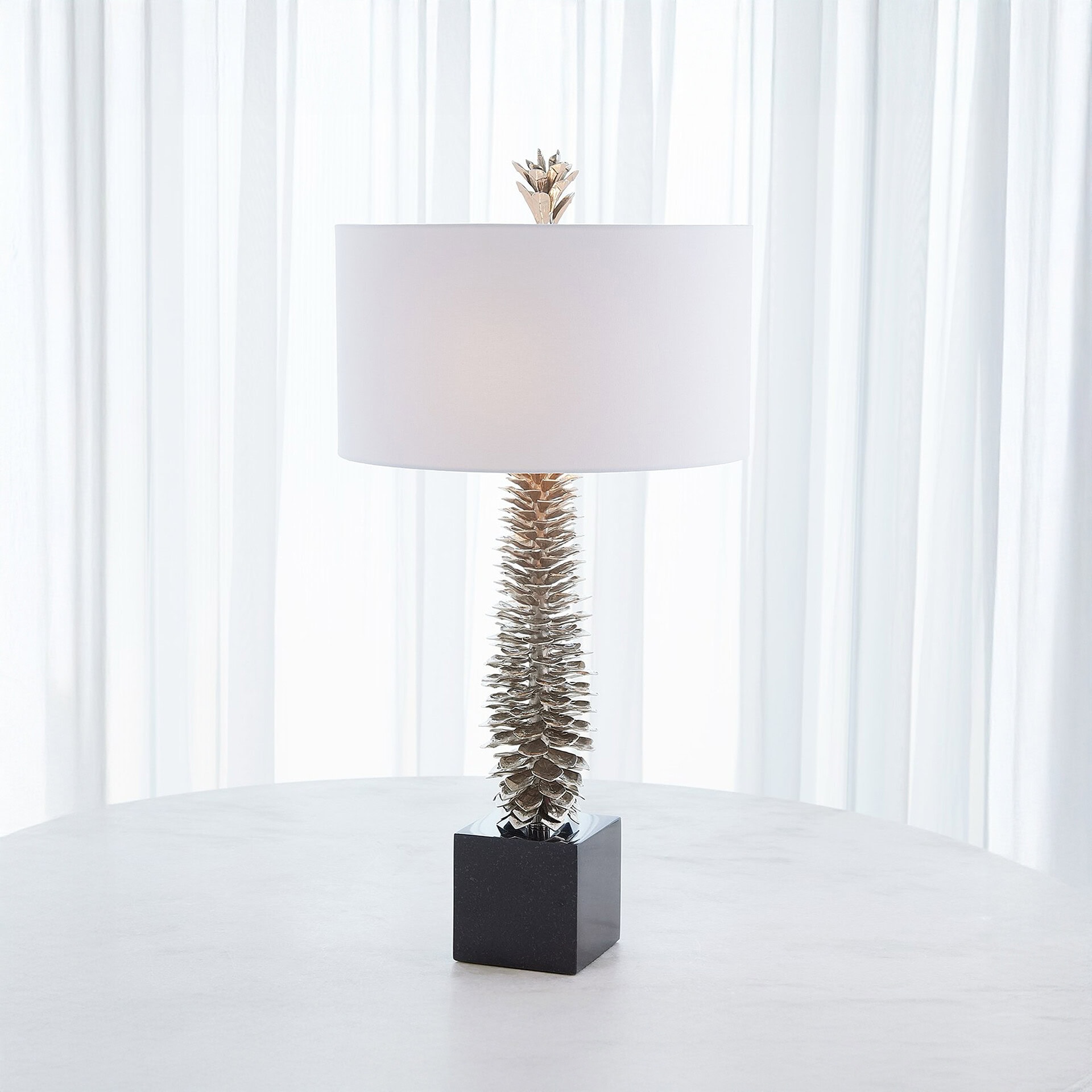 A modern table lamp with a white cylindrical shade, a silver-toned textured stem resembling a pinecone, and a black square marble base, on a white table in front of sheer white curtains.