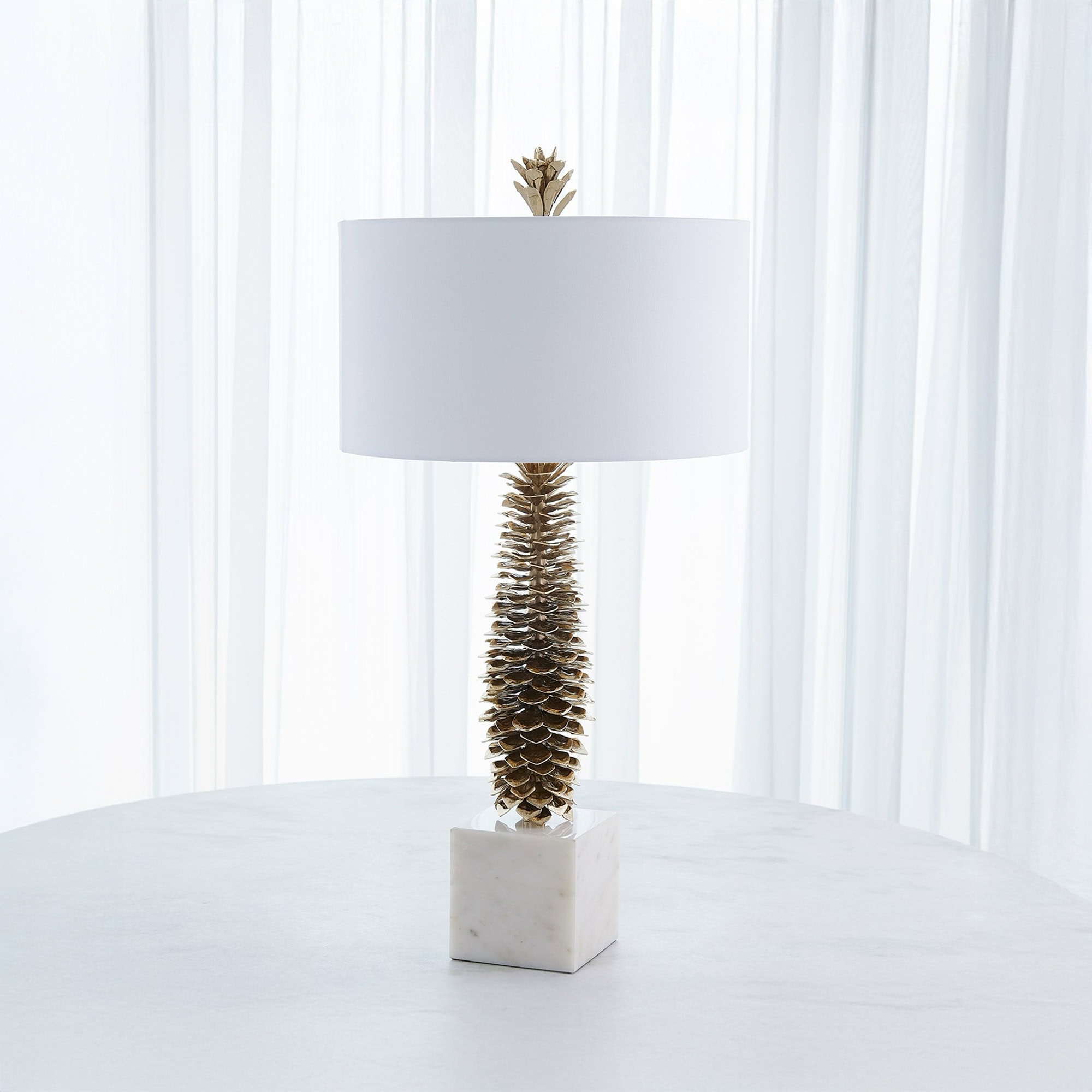 A gold pinecone-textured table lamp with a white drum shade and a square white marble base, sitting on a round marble table in front of sheer white curtains.