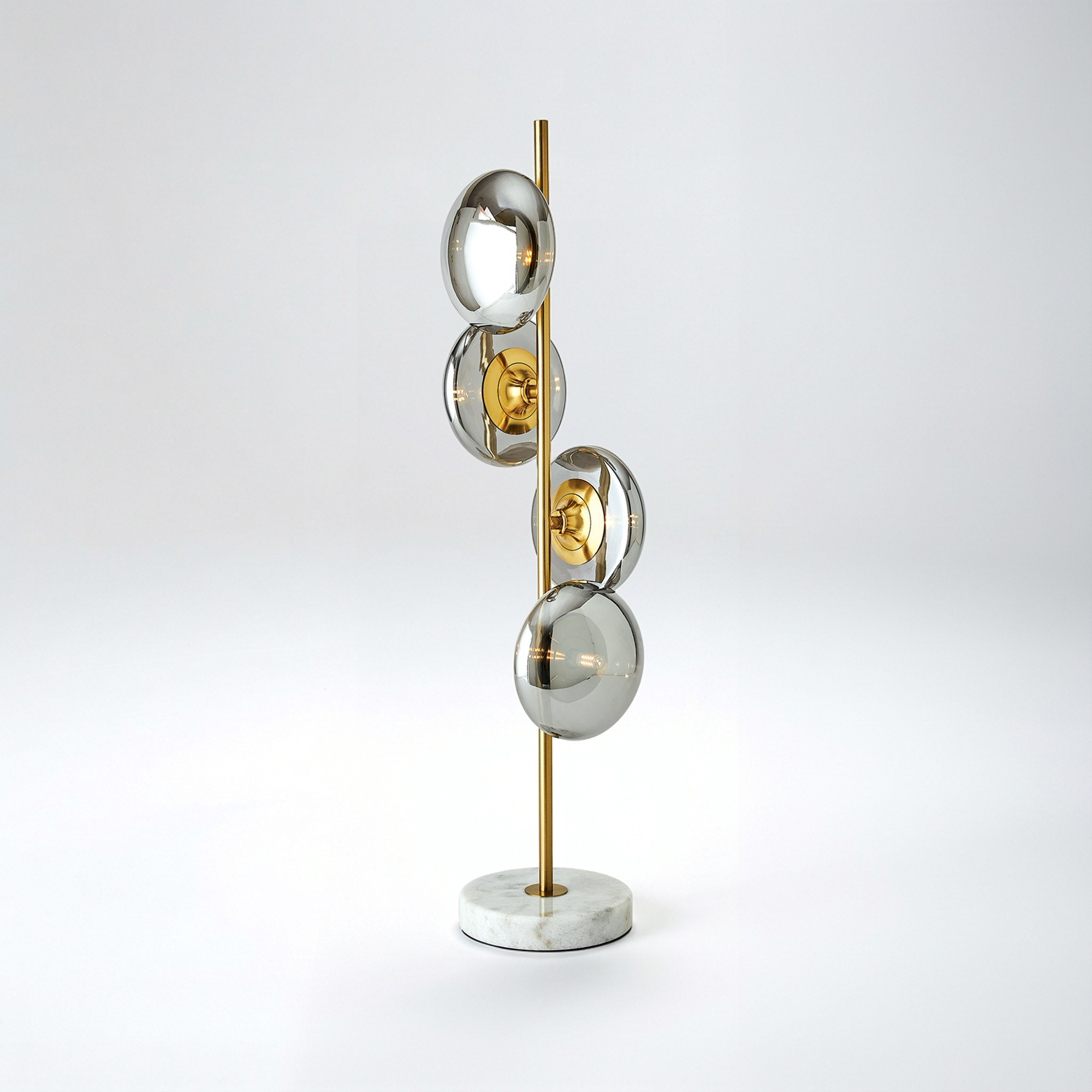 A modern table lamp with a round white marble base, a slender brass stem, and four staggered smoked glass globes, each containing a filament bulb, set against a light grey background.