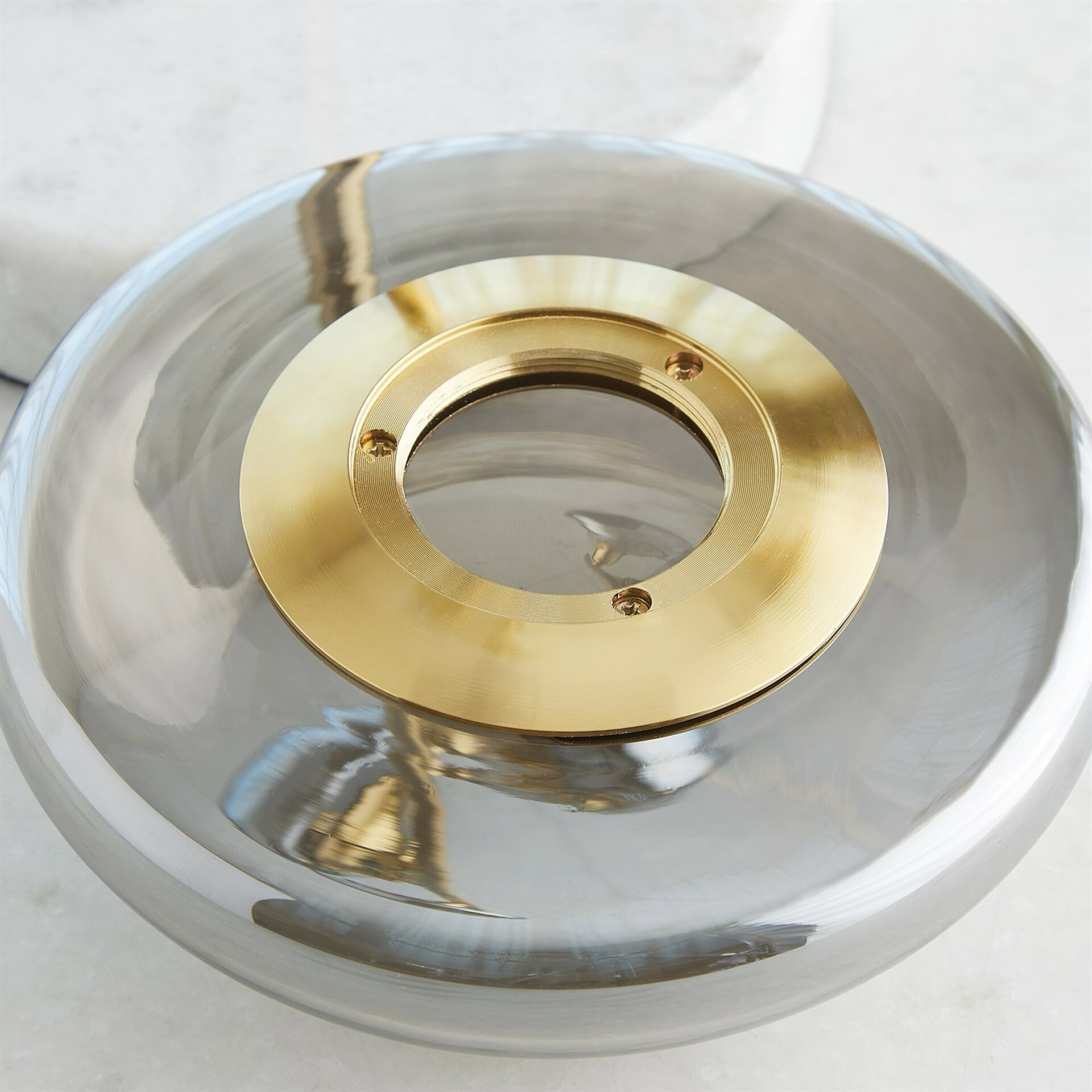 Top-down view of a circular, smoky gray glass object with a central brushed brass ring secured by three screws. The glass is highly reflective.