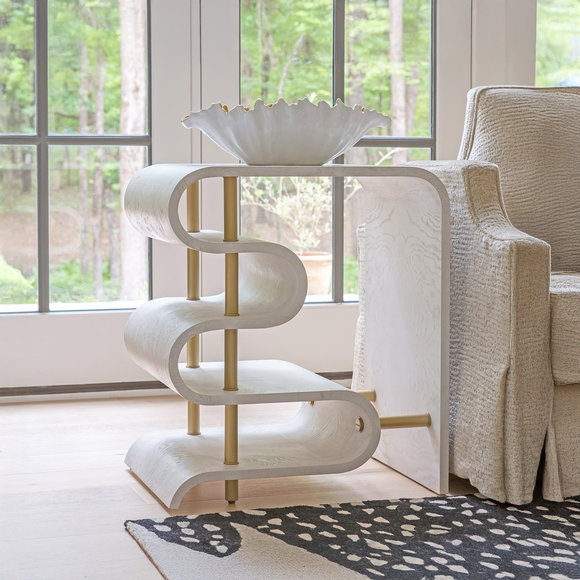 A modern white S-curve side table with brass accents, holding a white ruffled bowl with a gold rim. A beige armchair, light wood floor, and a black and white patterned rug are visible, with green trees outside a large window in the background.
