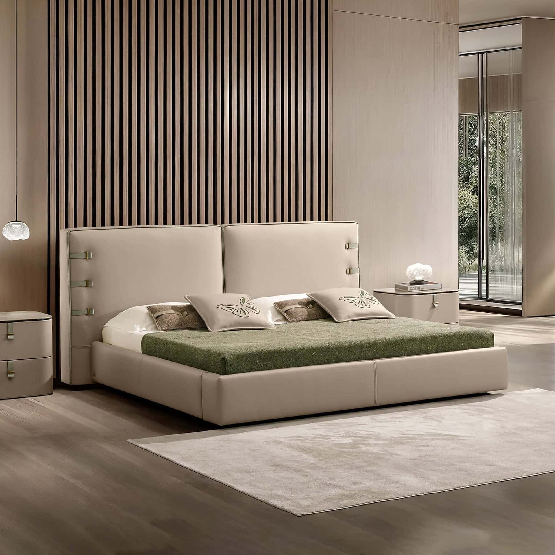 A modern bedroom with a beige upholstered bed, green blanket, and butterfly pillows. A slatted wooden accent wall is behind the bed, and a large window shows green trees. Beige nightstands with lamps flank the bed.