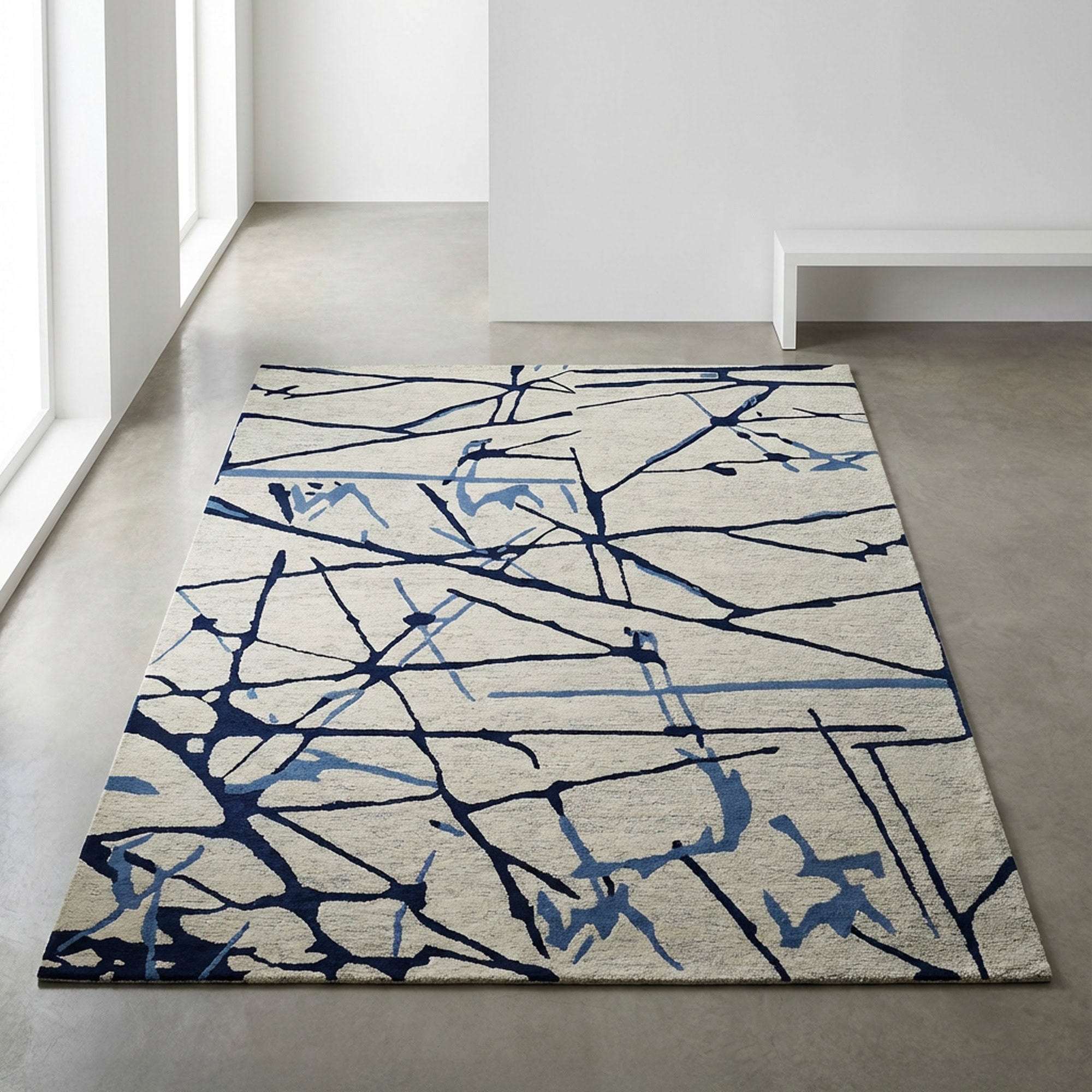 A large rectangular area rug with an abstract pattern of dark and light blue lines and shapes on a light background, placed on a concrete floor in a modern, minimalist room with white walls and windows.
