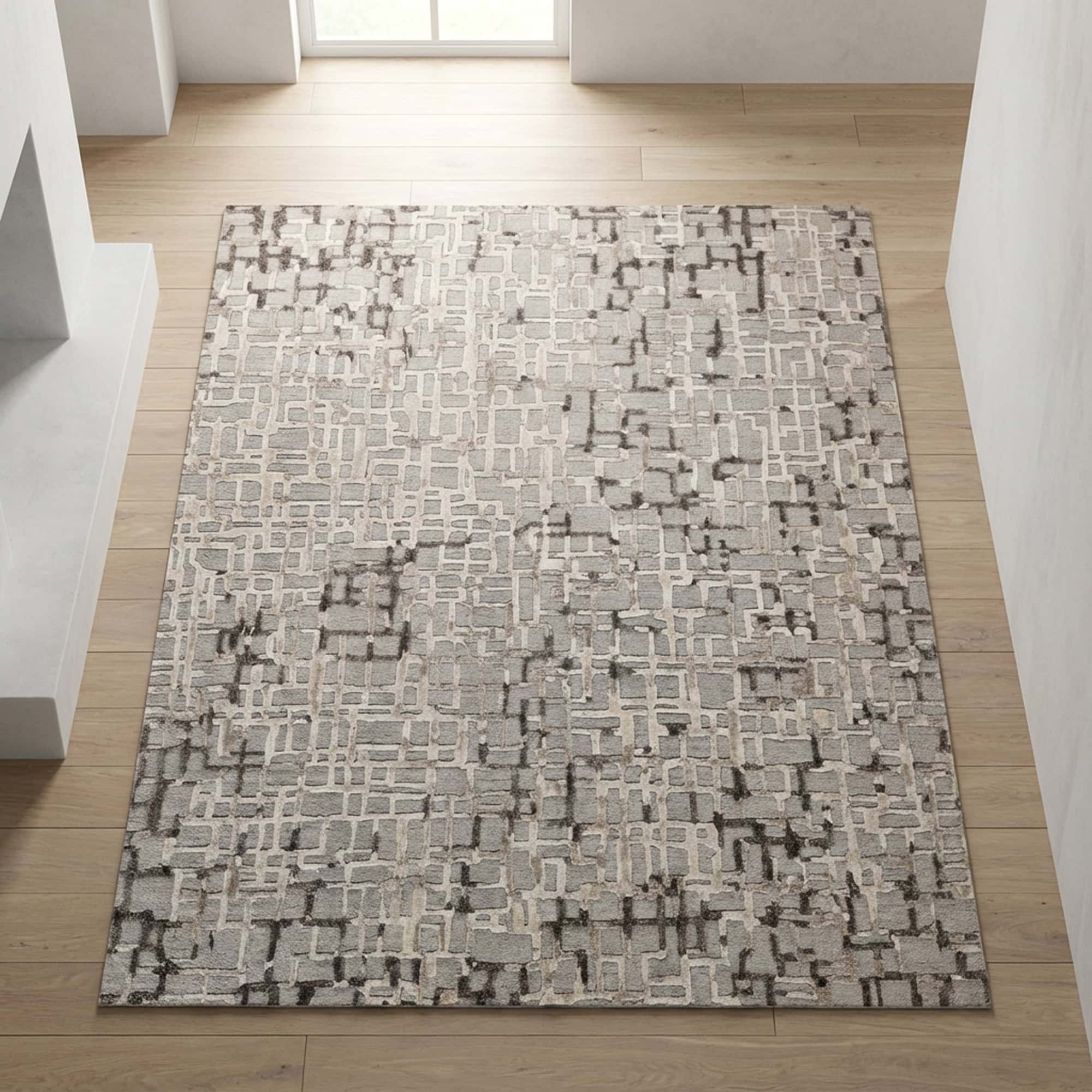 A large rectangular area rug with an abstract grey, cream, and charcoal geometric pattern on a light hardwood floor in a bright, modern living room. A beige sofa, wooden side table, and lamp are in the background, with tall windows on the left.