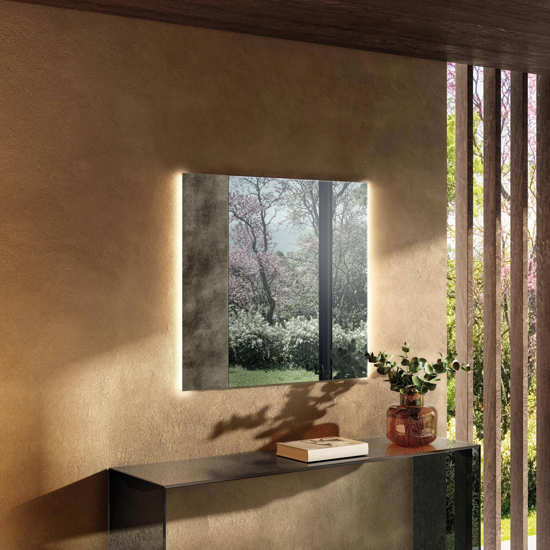 A backlit rectangular mirror on a textured brown wall reflects a vibrant outdoor scene with trees and blossoms. Below it, a dark console table holds a book and a vase with green foliage. A slatted wooden window on the right reveals more of the sunny outdoor landscape.