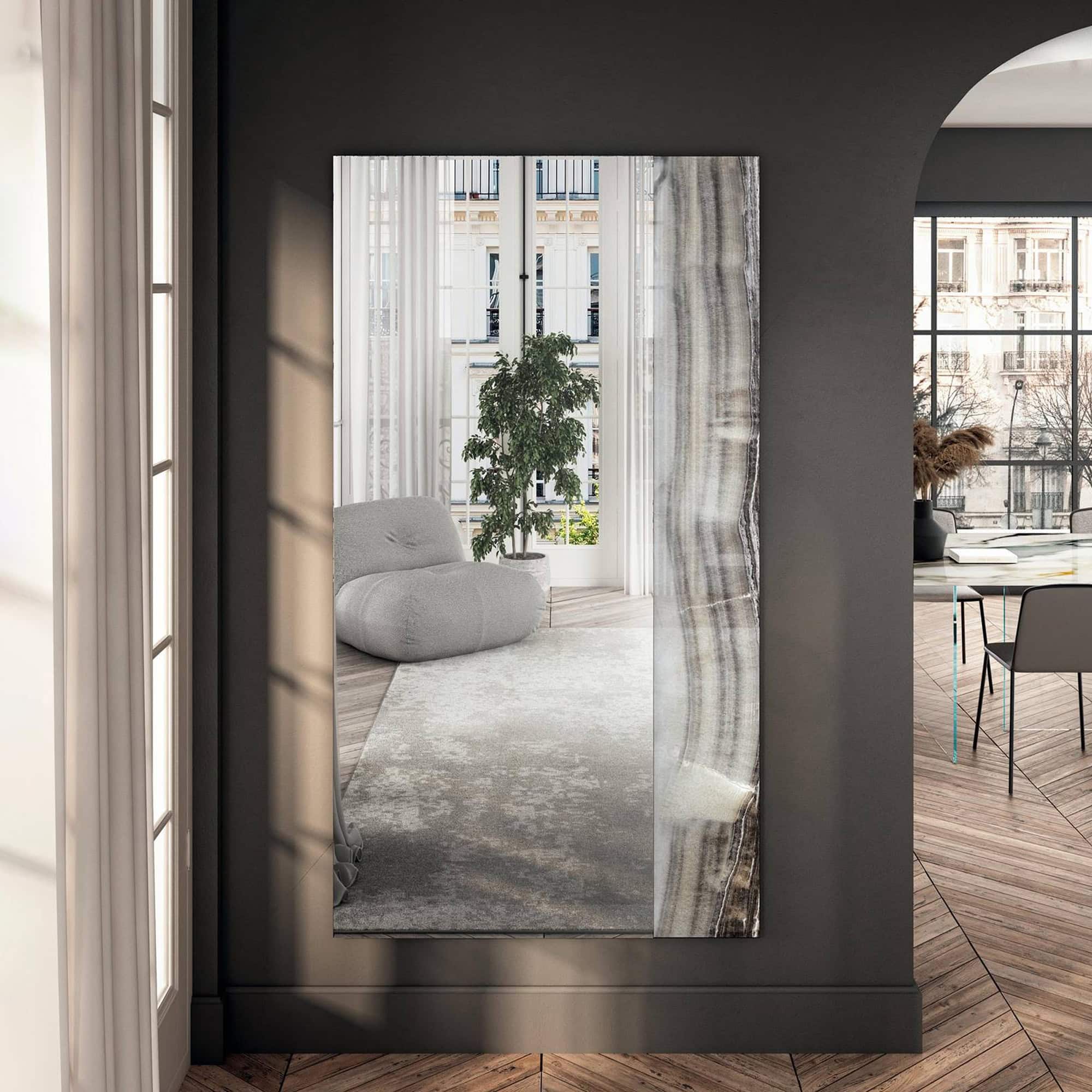 A large, rectangular full-length mirror with a natural stone-patterned edge leans against a dark gray wall, reflecting a bright room with a gray armchair, potted plant, and a city view.