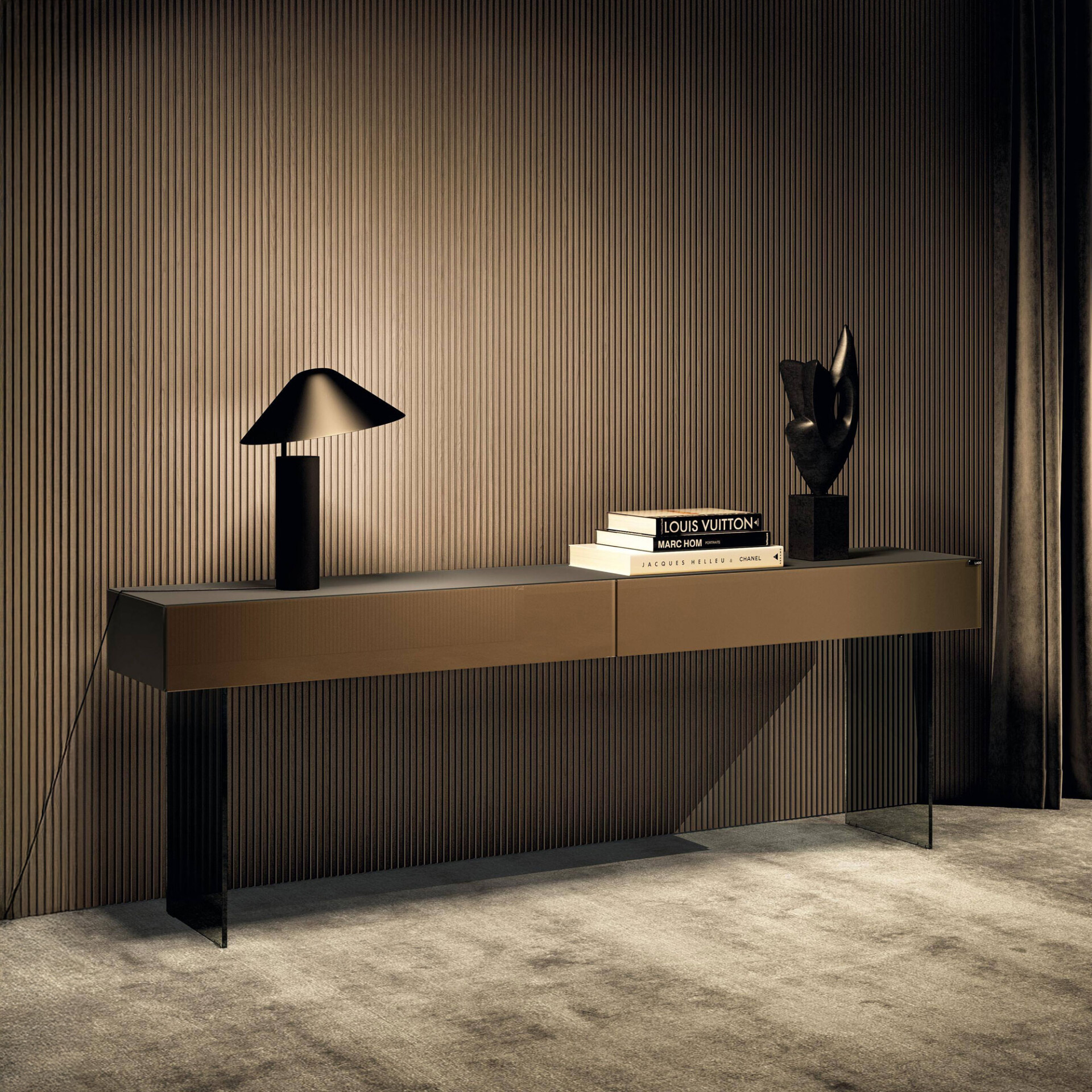 A modern console table with a black lamp, stacked books, and an abstract sculpture against a textured wood-slat wall and dark curtain.