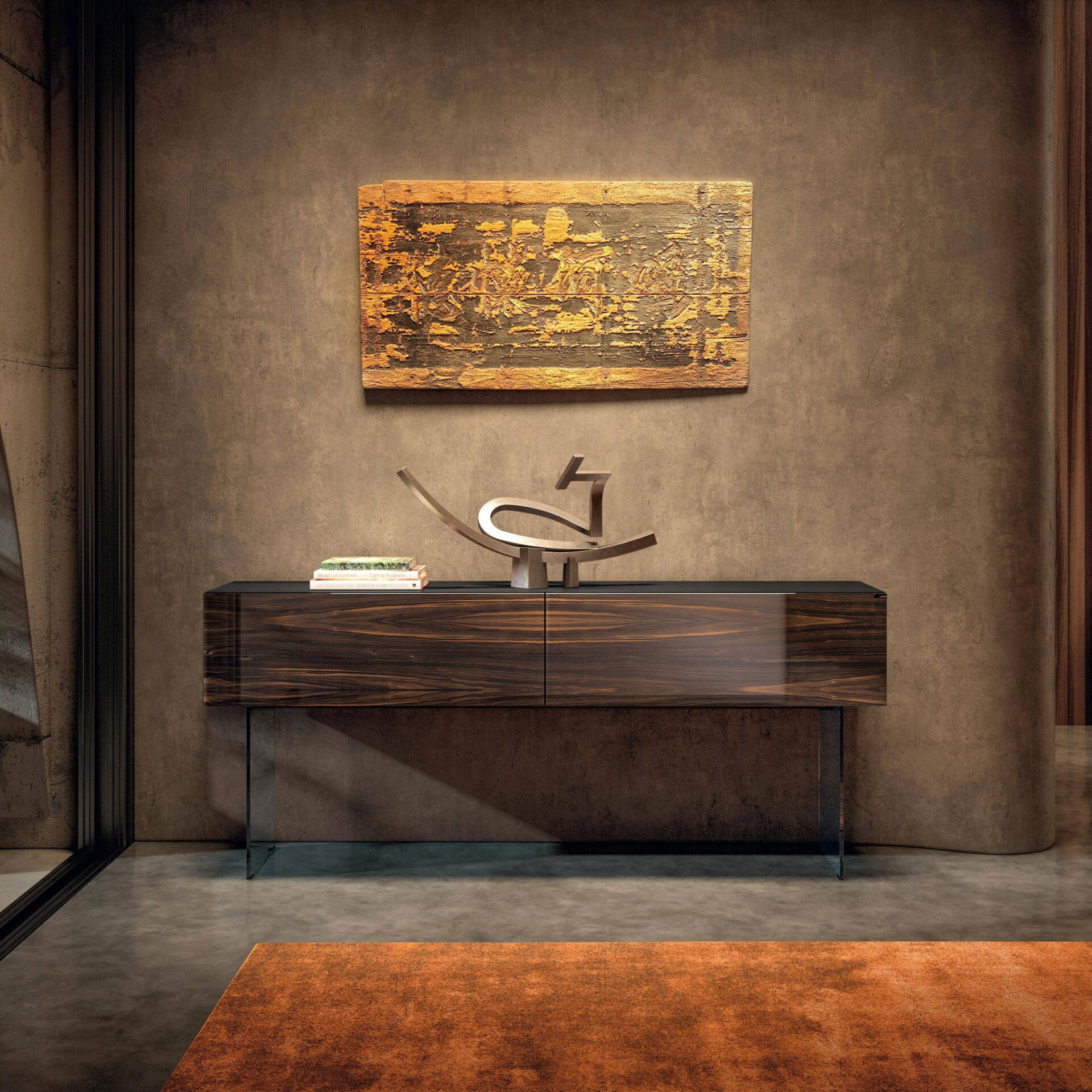 A modern interior with a dark wood console table, an abstract metallic sculpture, and a stack of books. Above it hangs a textured, golden-orange and dark brown wall art piece. The room features warm brown textured walls, a grey concrete floor, and an orange-rust rug.