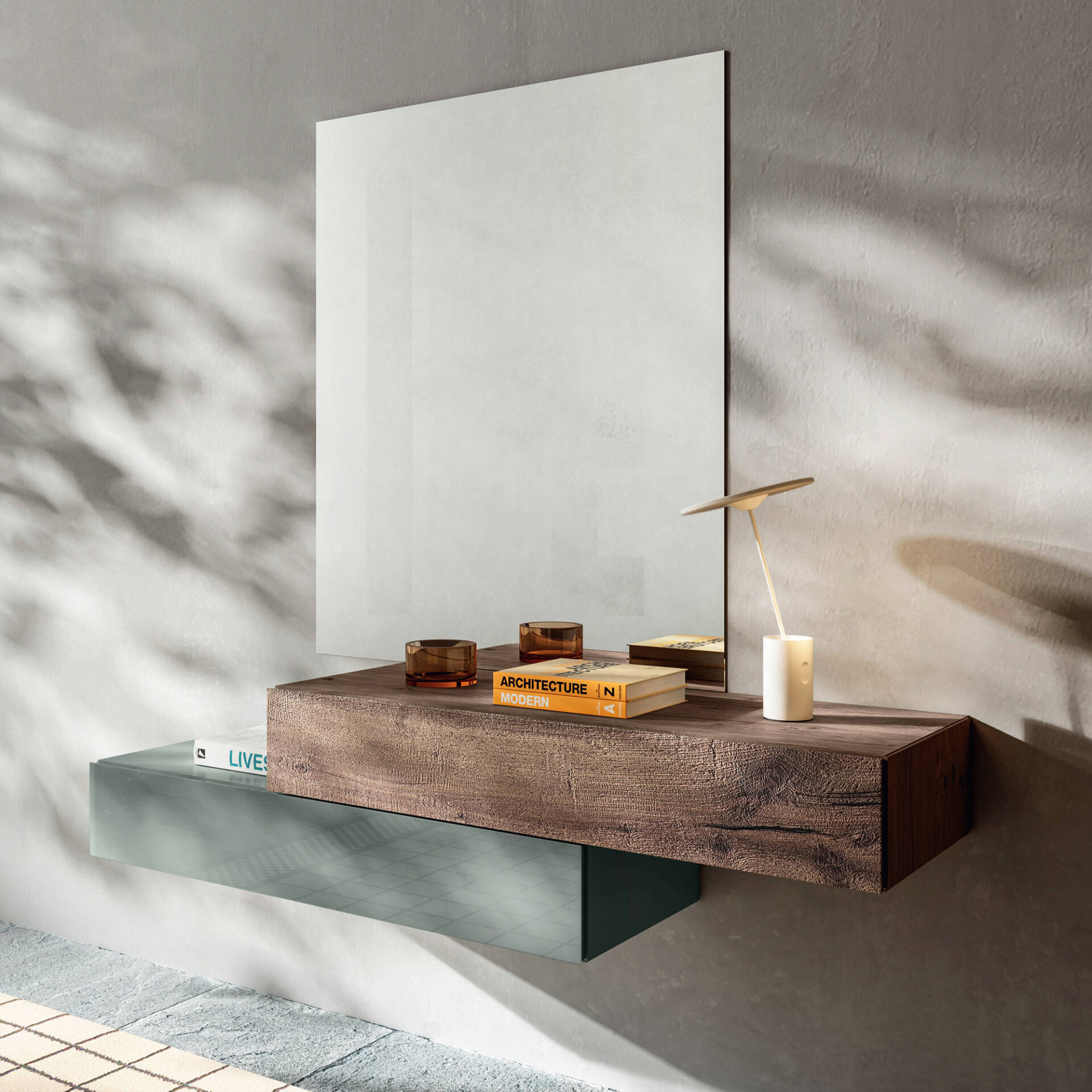 A modern entryway setup featuring a large rectangular mirror, a floating console with a textured wooden top and a light green glass lower shelf. The console is decorated with amber glass votives, books, and a minimalist table lamp, all set against a textured gray wall with dappled shadows.