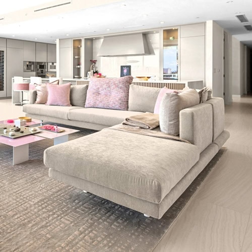 A modern open-concept living room featuring a large L-shaped beige sectional sofa with pink throw pillows, a pink and white coffee table, and a contemporary light-colored kitchen in the background.