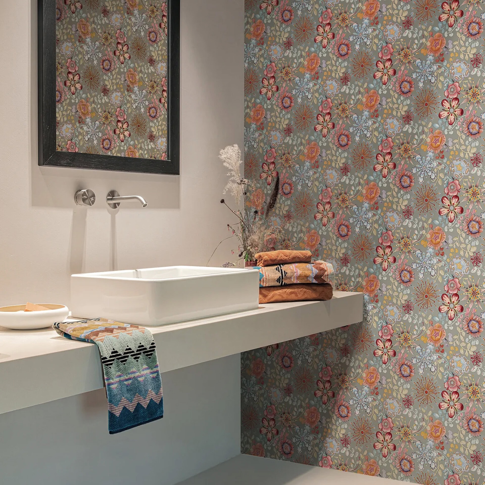 A modern bathroom with a white vessel sink on a floating counter, a black-framed mirror, and a wall covered in vibrant floral wallpaper. Colorful patterned towels and dried plants add decorative accents.