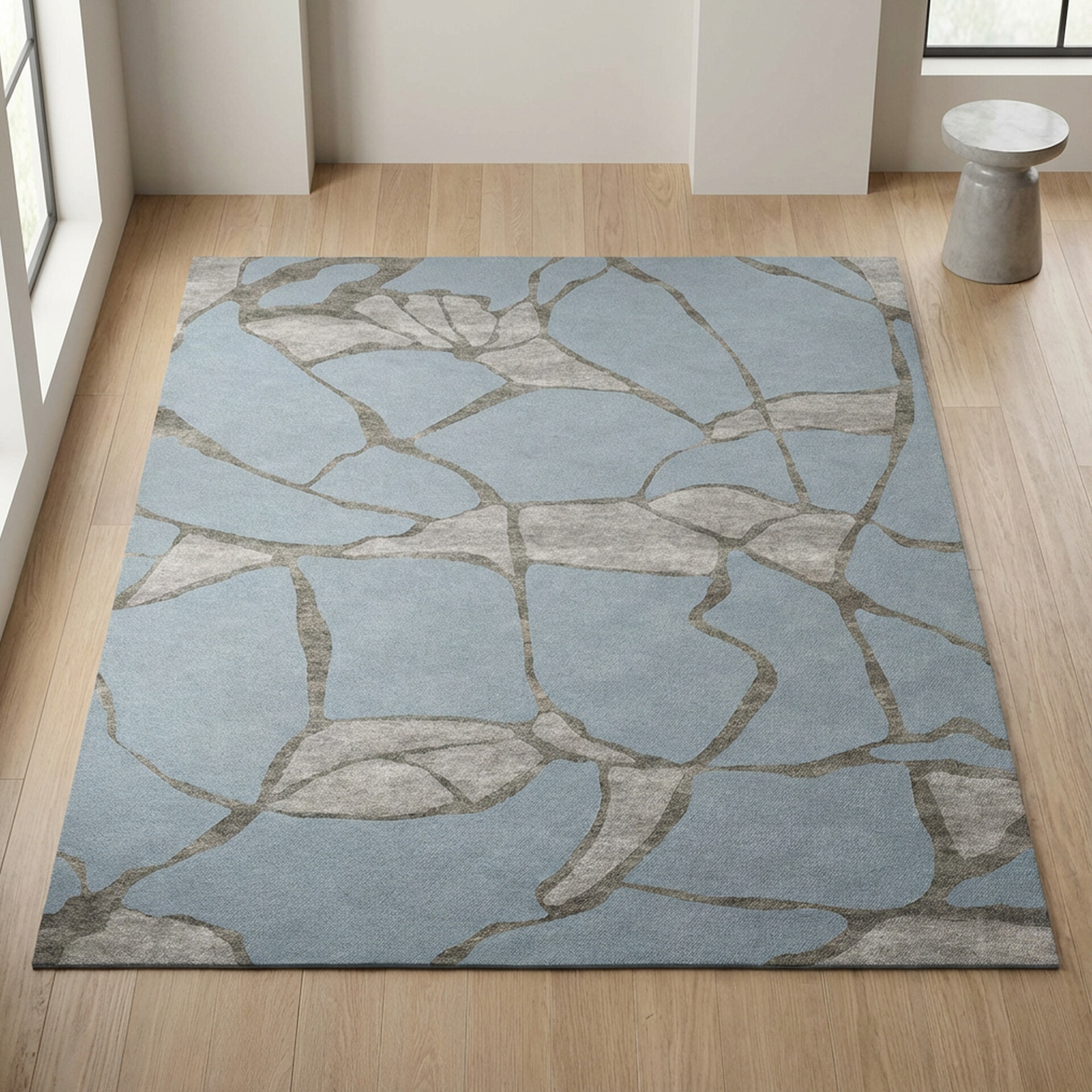 A light blue and grey abstract area rug with an organic, cracked-ice pattern on a wooden floor in a modern room with a window and a grey stool.